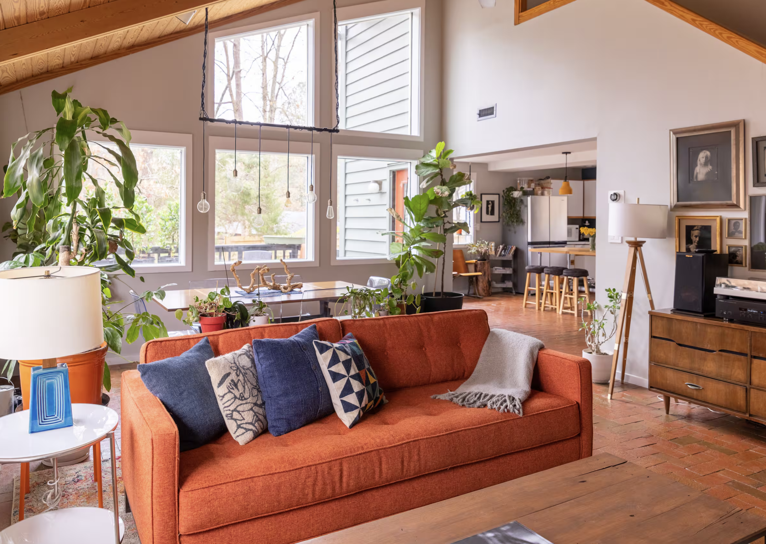 A cozy living room with a red-orange sofa adorned with patterned throw pillows, a white side table with a blue holder and a white lamp, a large green plant in a black pot, and a wall decorated with framed black and white portraits. The room has large