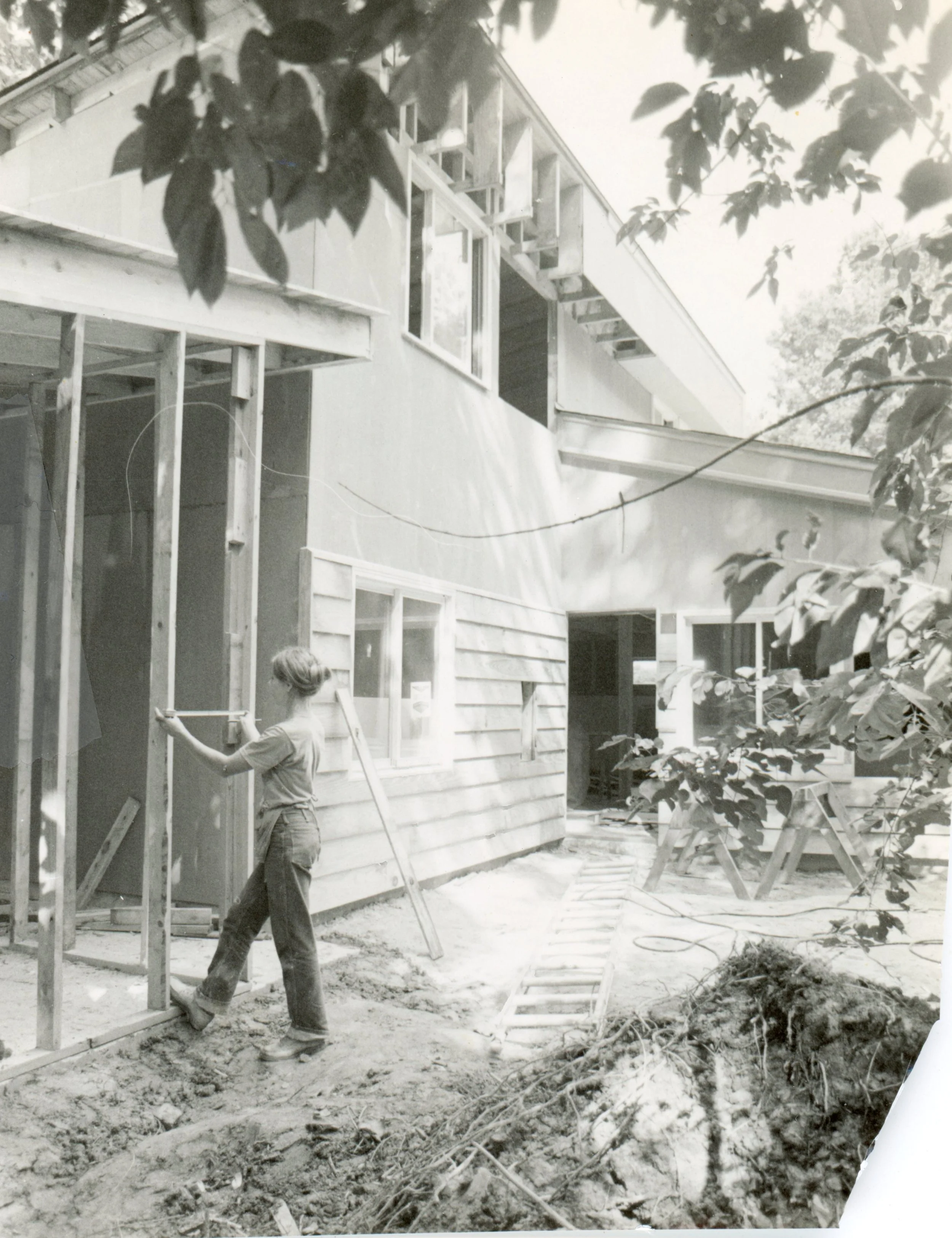 Black and white photo of a house under construction with a woman in casual clothes and boots working on the frame, surrounded by construction materials and tools.