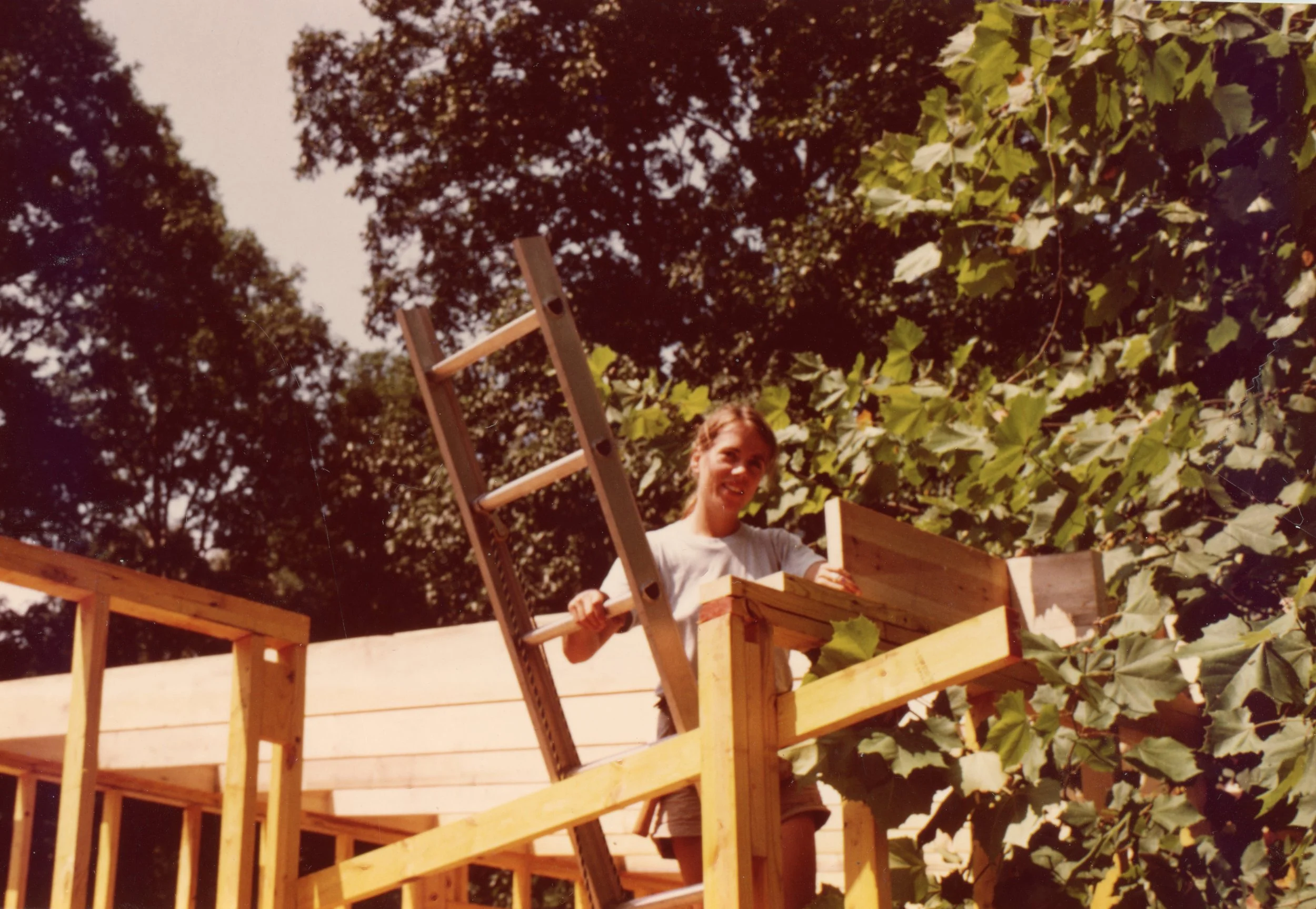 A person standing on a wooden construction site, smiling, holding onto a ladder, with trees and leaves in the background.