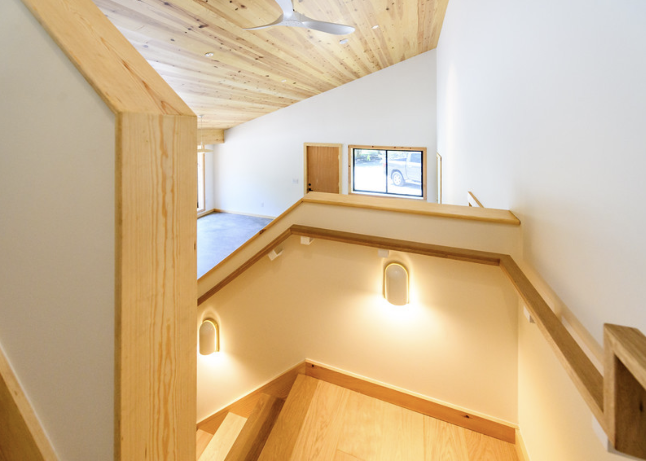 Interior view of a staircase with wooden handrails, stairs, and ceiling, with windows allowing natural light and a car visible outside.