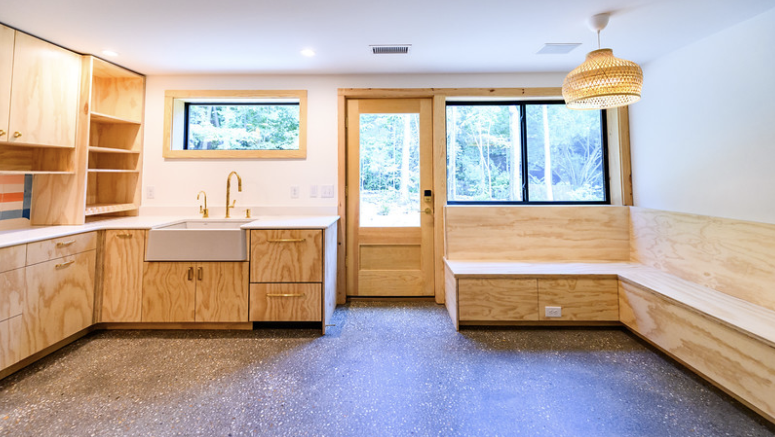 Interior of a modern kitchen with light wood cabinets, a white farmhouse sink, a window and a glass door. A corner bench is built with matching wood. The ceiling has a wicker pendant light and recessed lighting. Outside, trees are visible through the