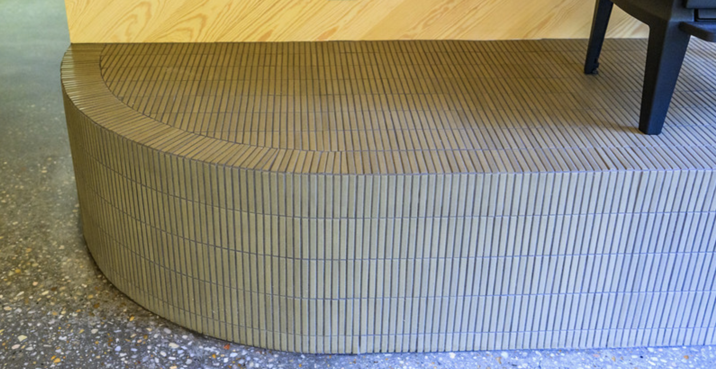 Close-up of a curved, tiled wooden bench with narrow, vertical black lines separating the tiles, part of a black chair leg is visible on the right, floor is speckled concrete.