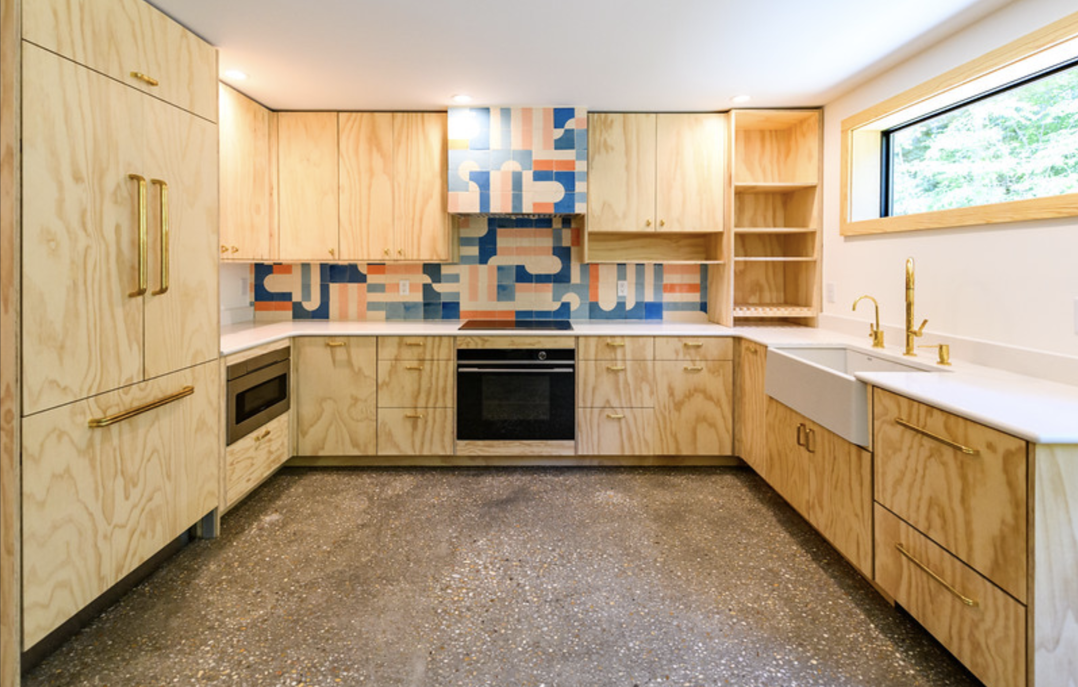 Kitchen with light wood cabinets, colorful geometric tiled backsplash, window with a view of greenery, gold faucet and handles, white countertop, and black oven.