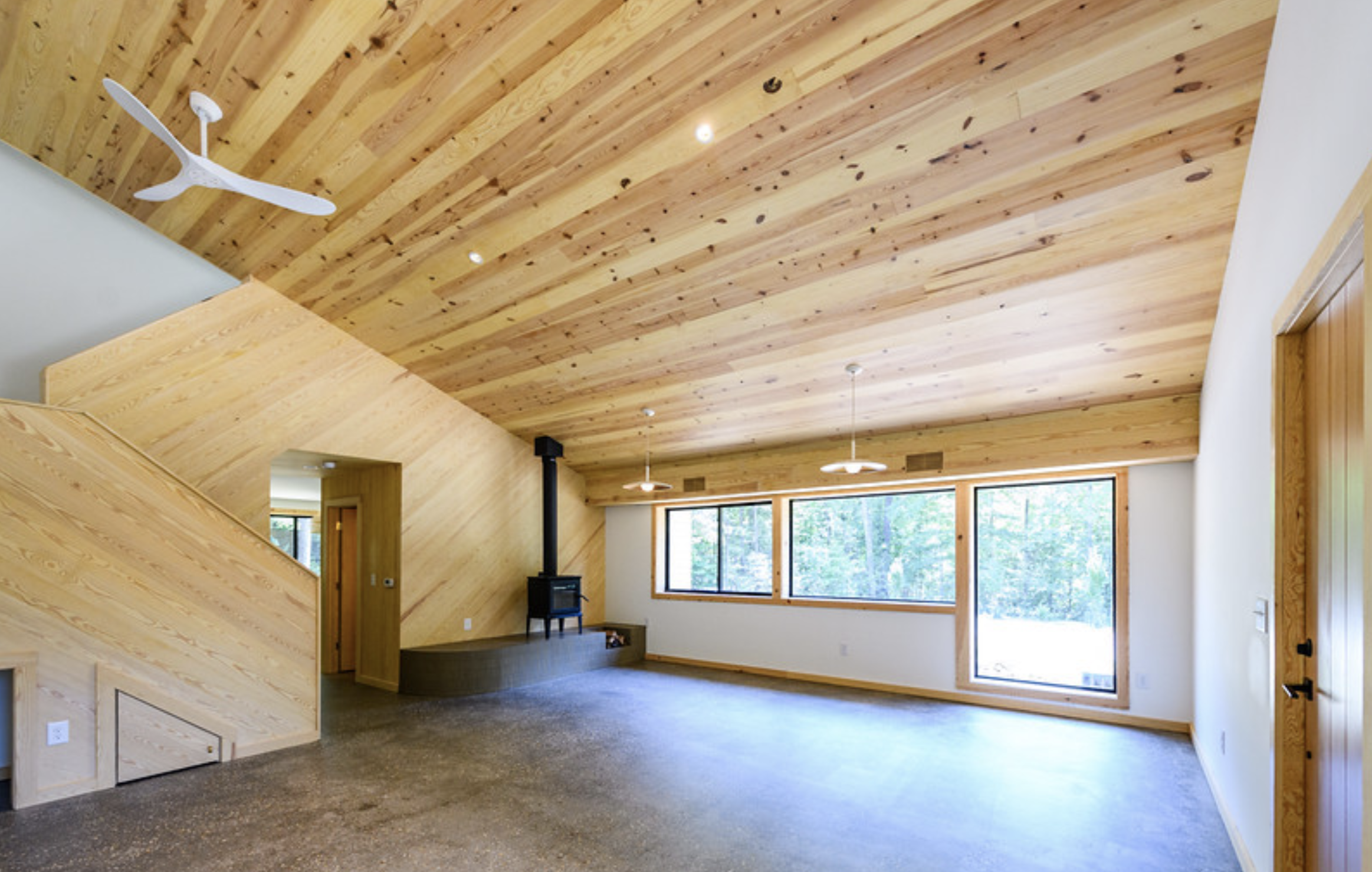 Interior of a room with a wooden vaulted ceiling, large windows letting in natural light, and a small stove with a pipe on the floor.