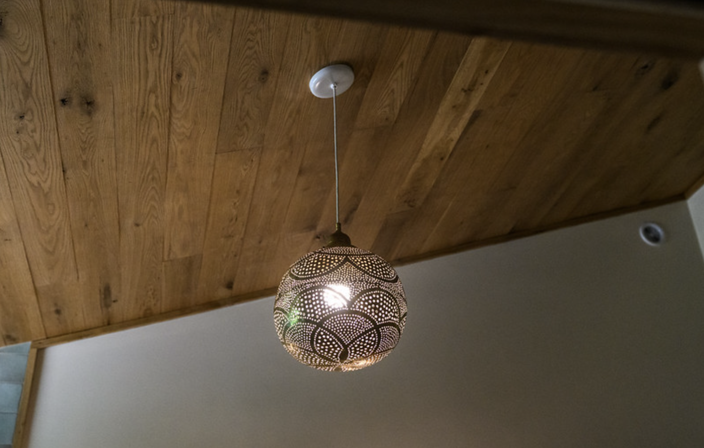 Decorative hanging pendant light with a intricate perforated pattern, hanging from a white ceiling mount, against a wooden ceiling and white wall.