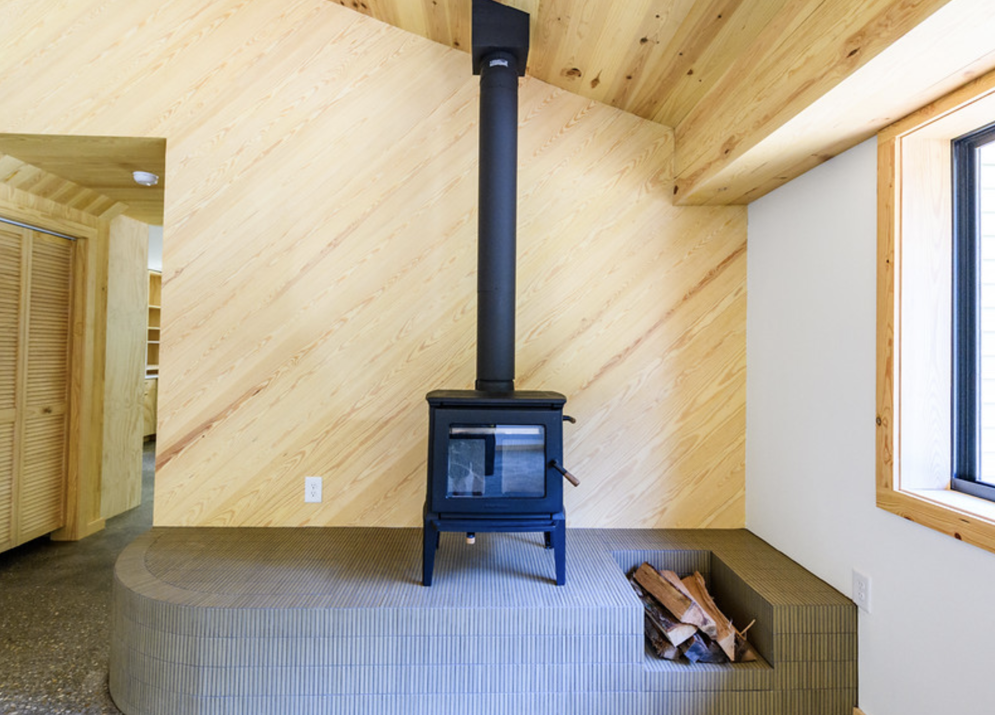 Interior of a room with a wood stove on a raised, gray tiled platform, wood logs nearby, a sloped wood-paneled ceiling, and a window on the right side.