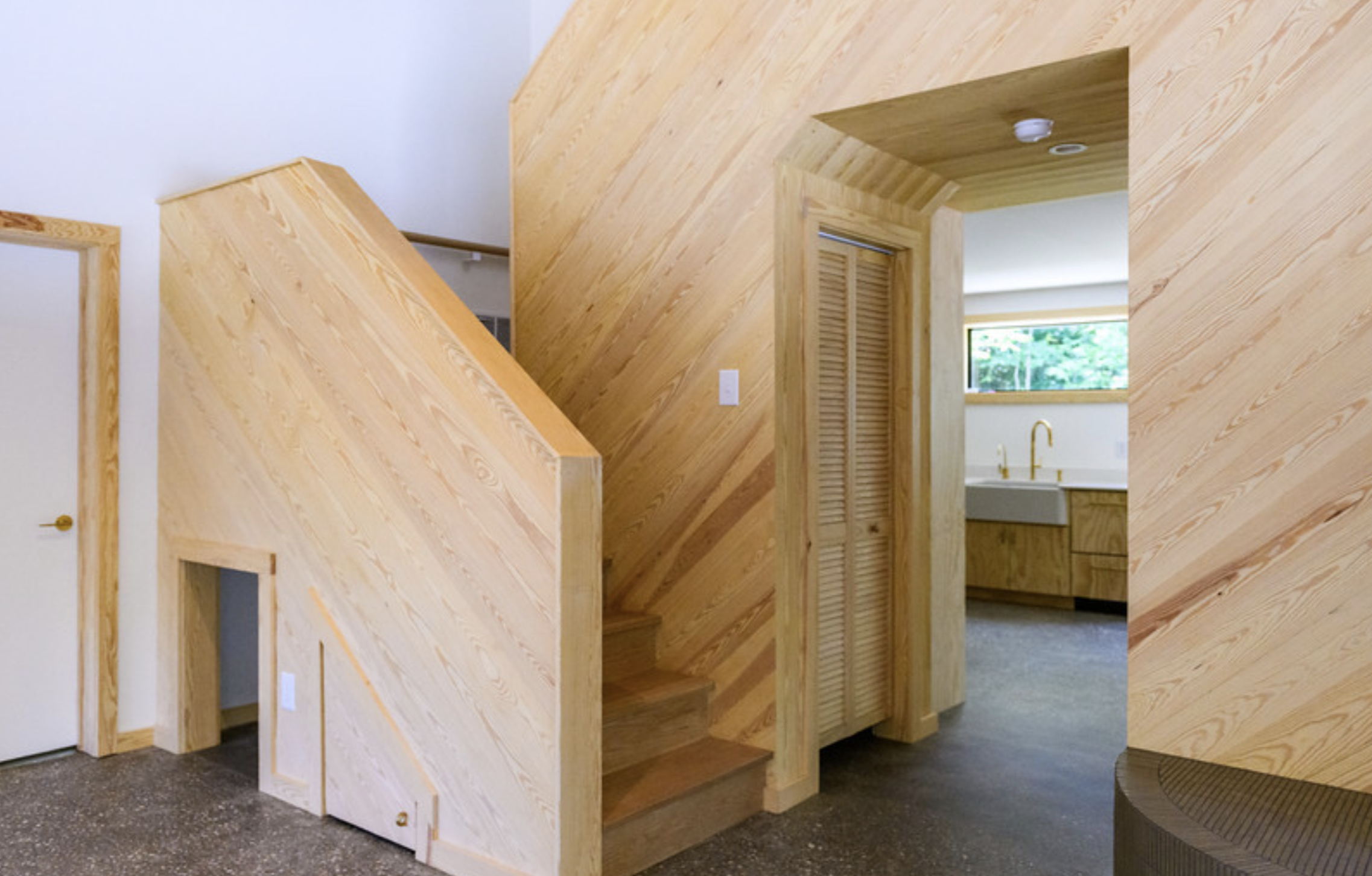 Interior view of a house with walls and ceiling finished in light-colored wood paneling, including a staircase with a wooden railing, a door with a louvered panel, a small window, and an opening leading to a kitchen with a window, white countertops, 