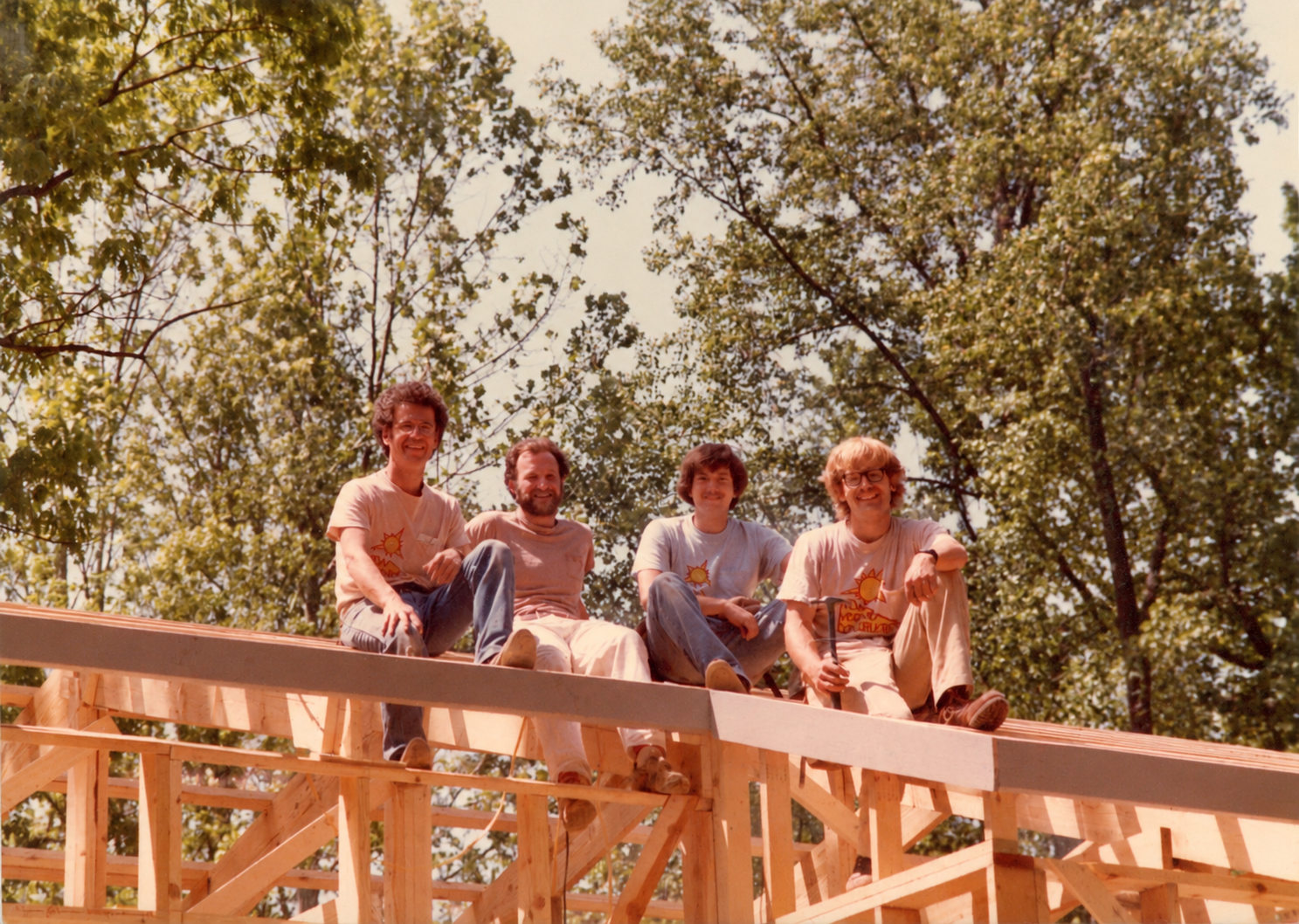 Four men sitting on a wooden construction framework outdoors with trees and a partly cloudy sky in the background.