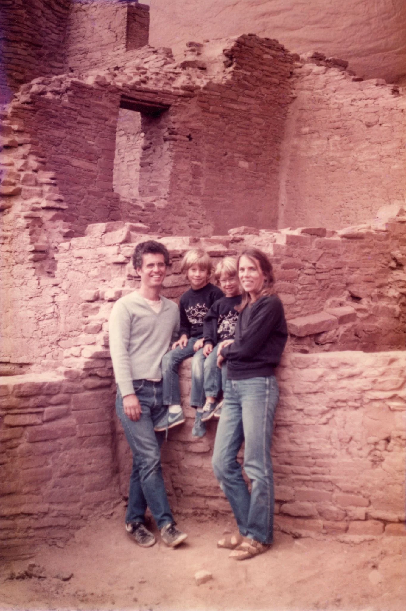 A family of four standing in front of ancient red brick ruins with layered walls. The man and woman are smiling, with two children sitting on the ruins between them. The background features weathered brick walls with an opening and remnants of an old structure.