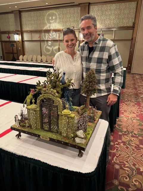 A smiling woman and man stand behind a detailed fantasy miniature landscape on a table at an indoor event.