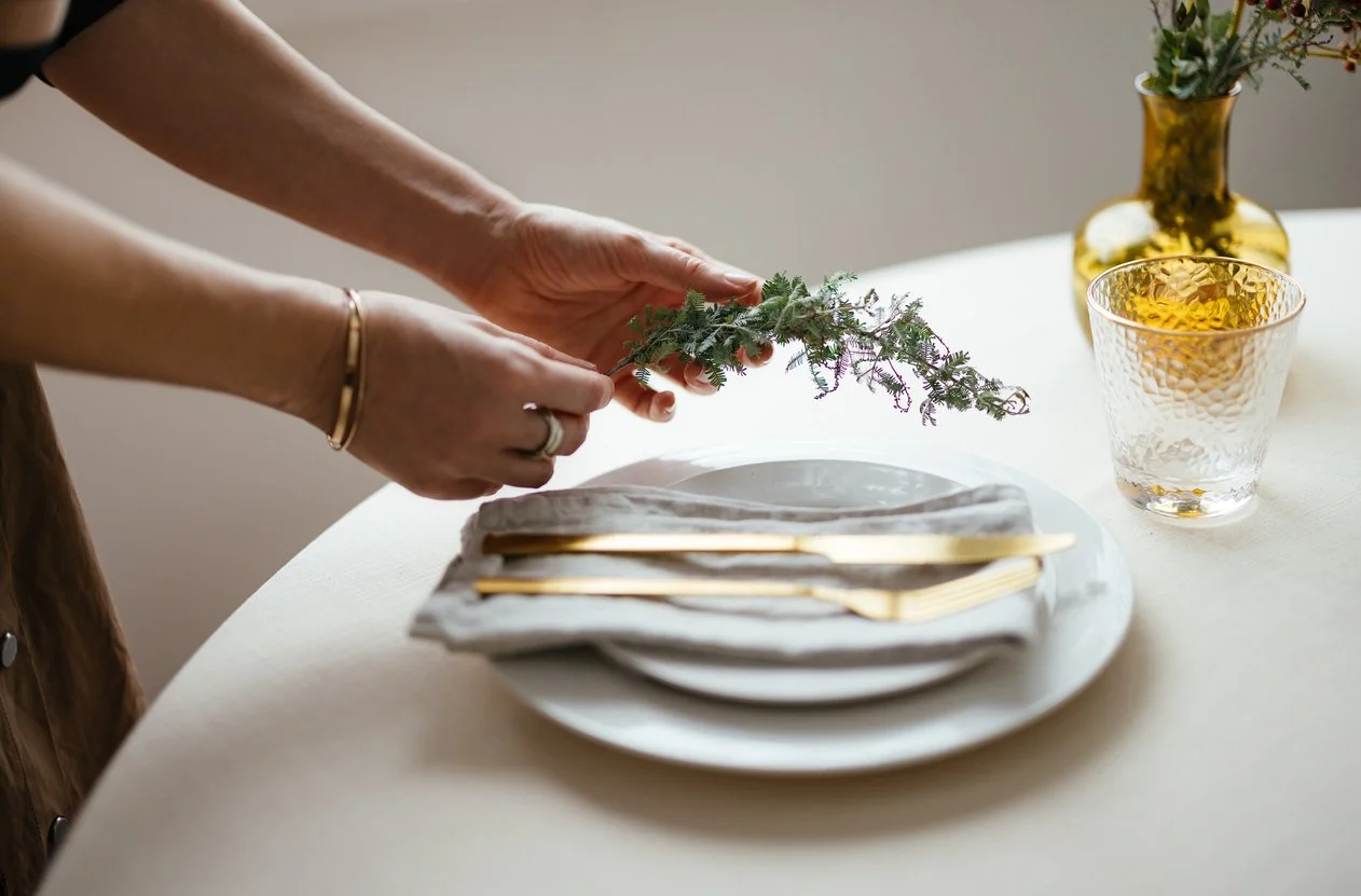 Person arranging a sprig of greenery on a table set with a white plate, gold cutlery, a cloth napkin, a patterned glass, and a gold vase with flowers.
