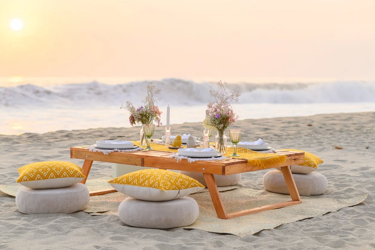 Beach picnic setup with a low wooden table, cushioned seats, and floral decorations during sunset.