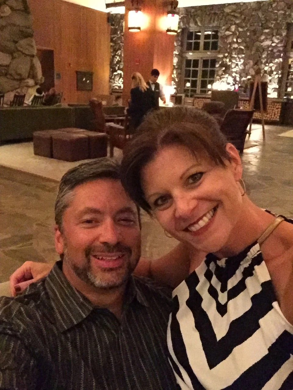 A smiling couple taking a selfie in a warmly lit hotel lobby with rustic stone and wood decor.