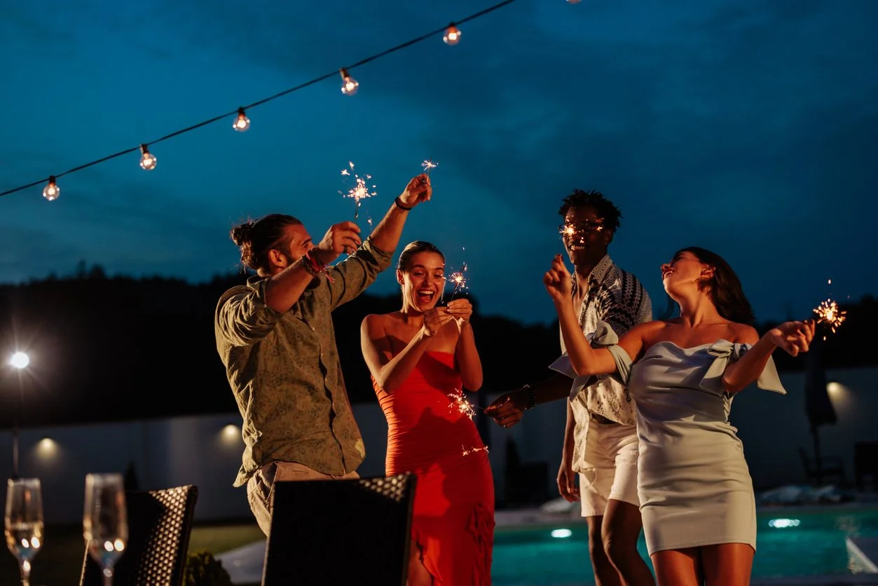 Four friends celebrating with sparklers outdoors at night, near a pool, under string lights.