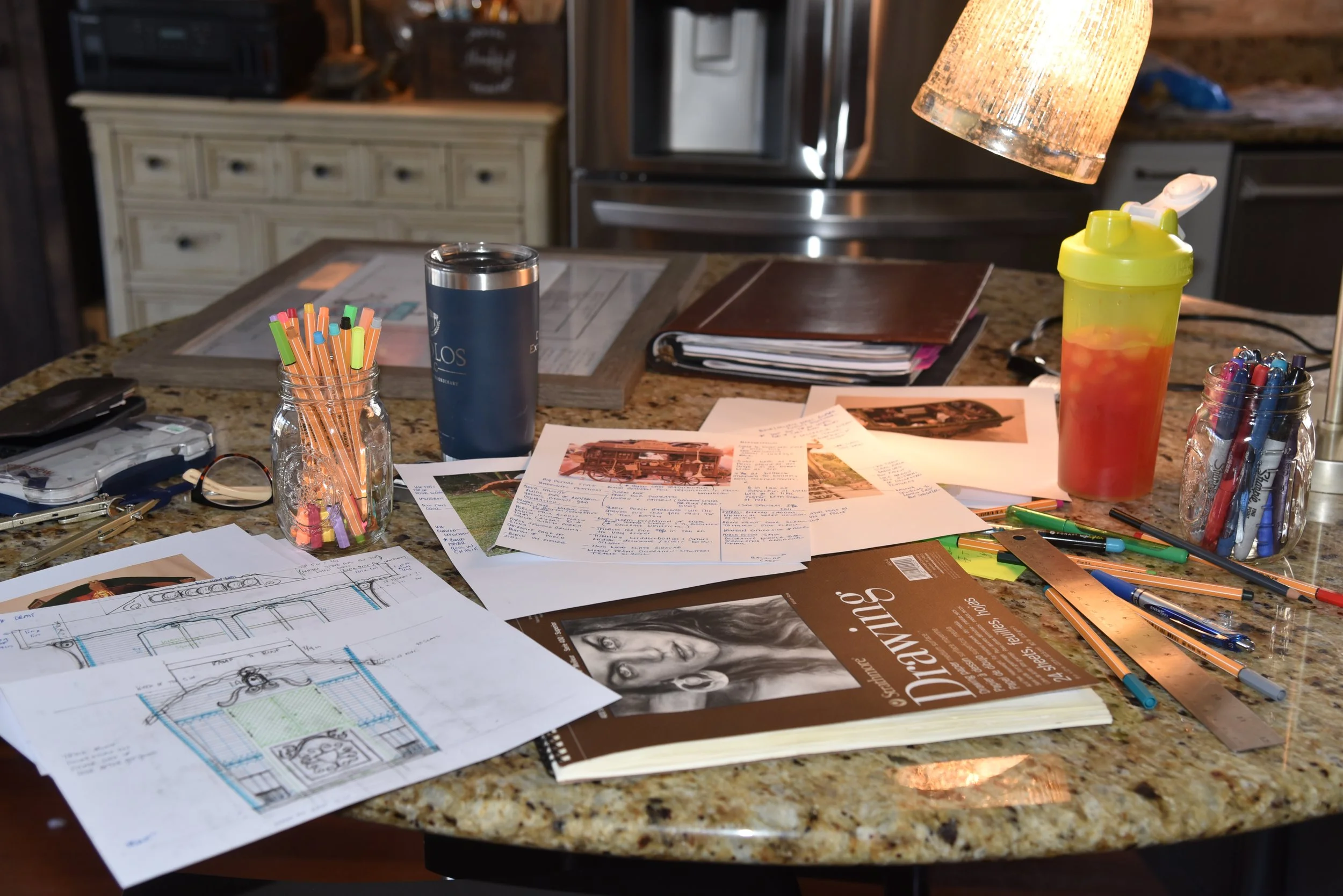 A cluttered kitchen countertop with papers, notebooks, pens, highlighters, a water bottle, a tumbler, and jars of colored pens. In the background, there is a lamp hanging over the counter.