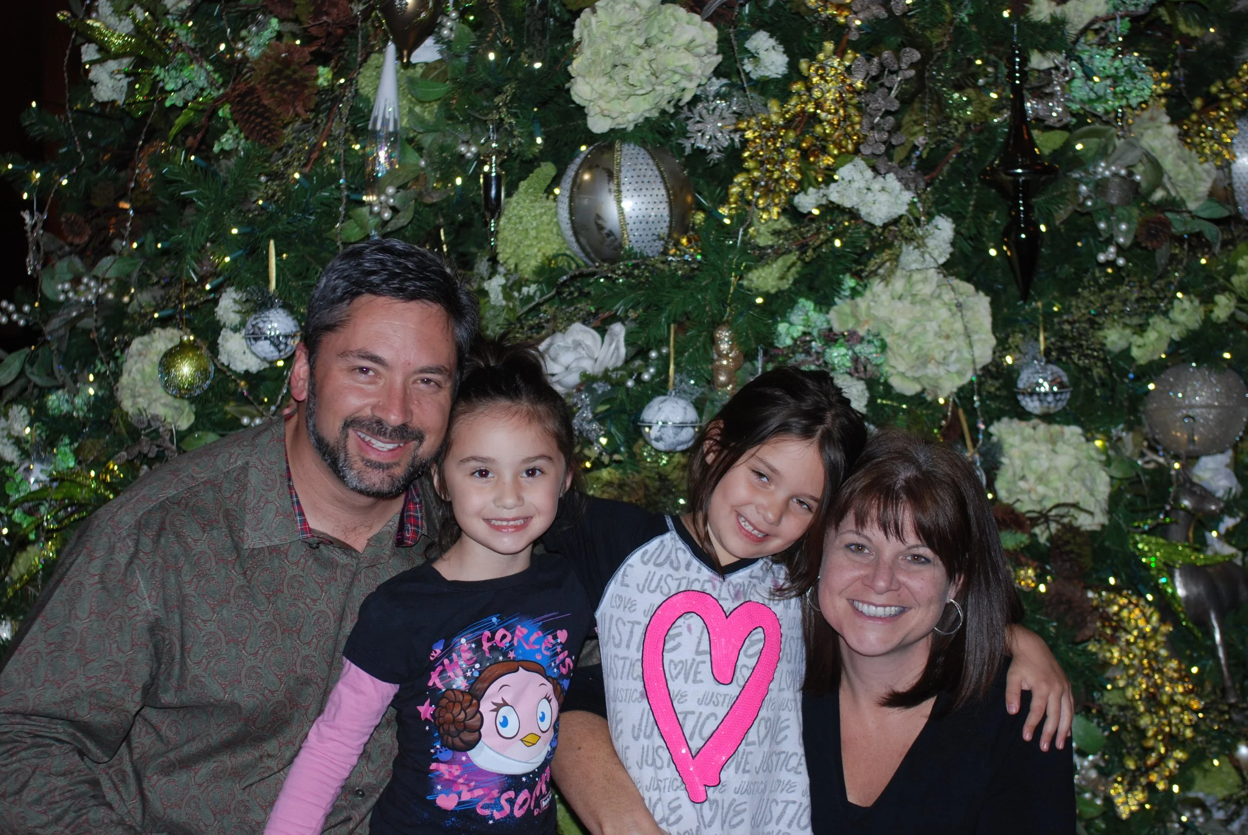 A group of five people, including two adults and three children, smiling and posing in front of a decorated Christmas tree with white and gold ornaments, lights, and flowers.