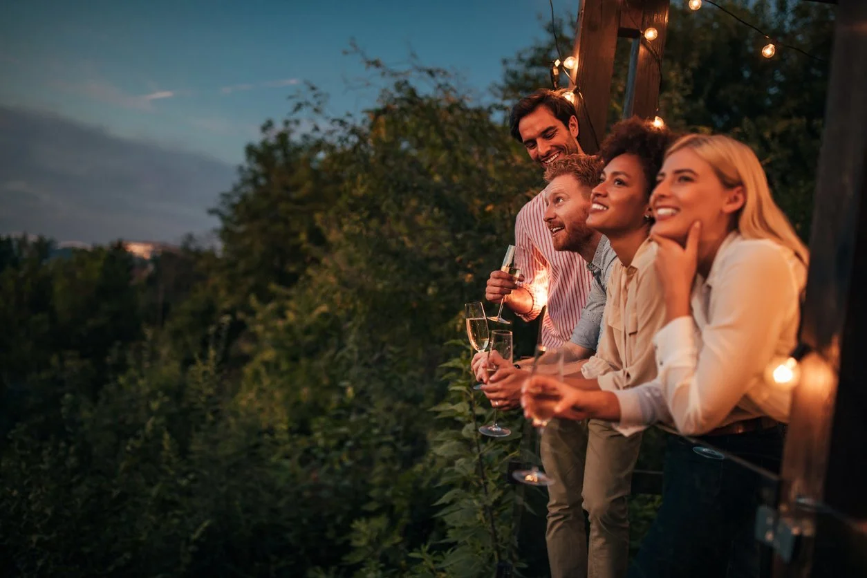 Group of four friends enjoying drinks on a balcony with string lights during sunset.