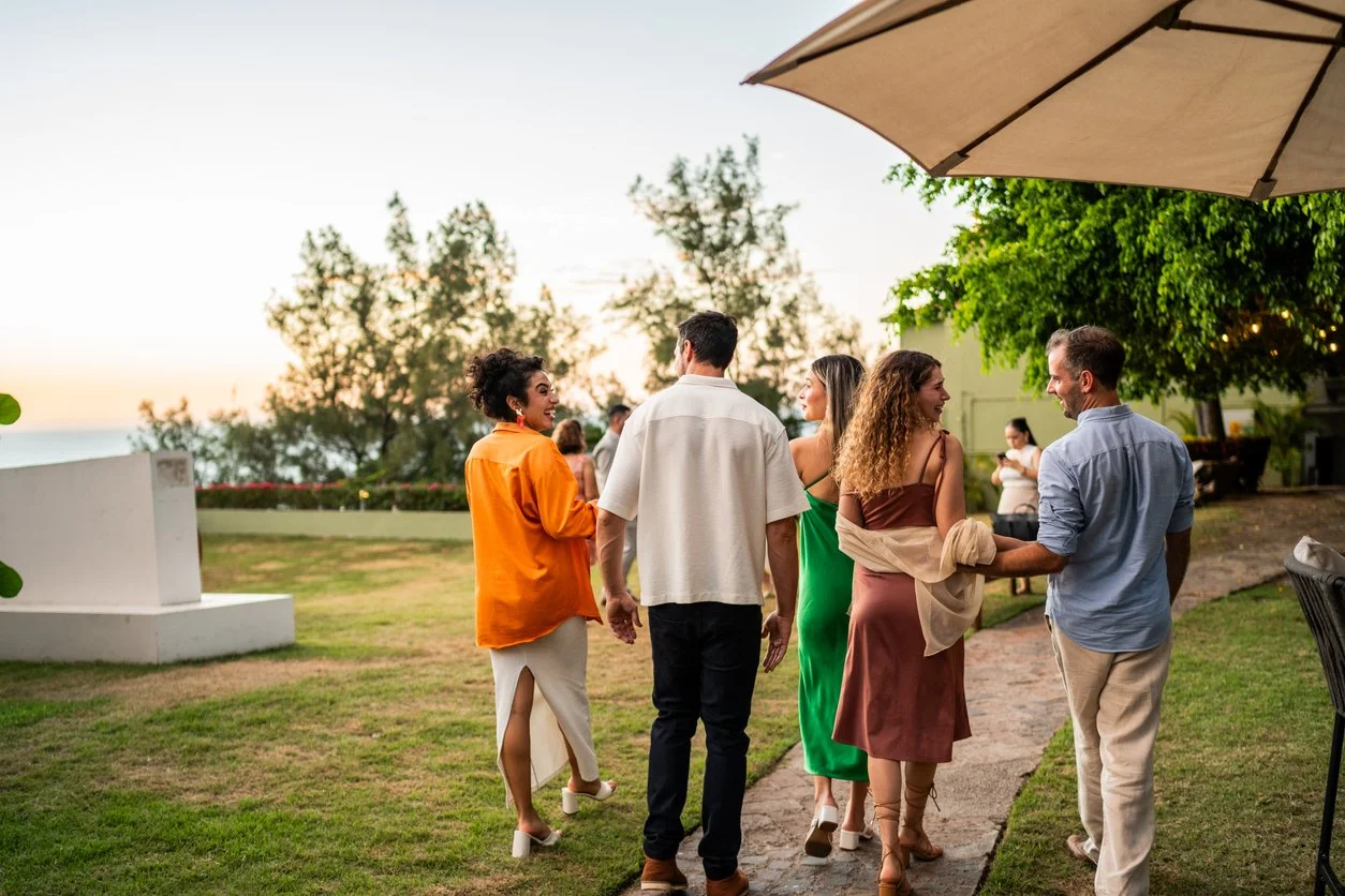 Group of people socializing outdoors during sunset, standing on a grassy area with trees and a white wall in the background.