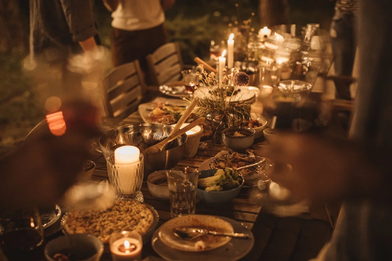 A dinner party outdoors at night with a long wooden table decorated with flowers and candles, surrounded by people, with various dishes and drinks on the table.