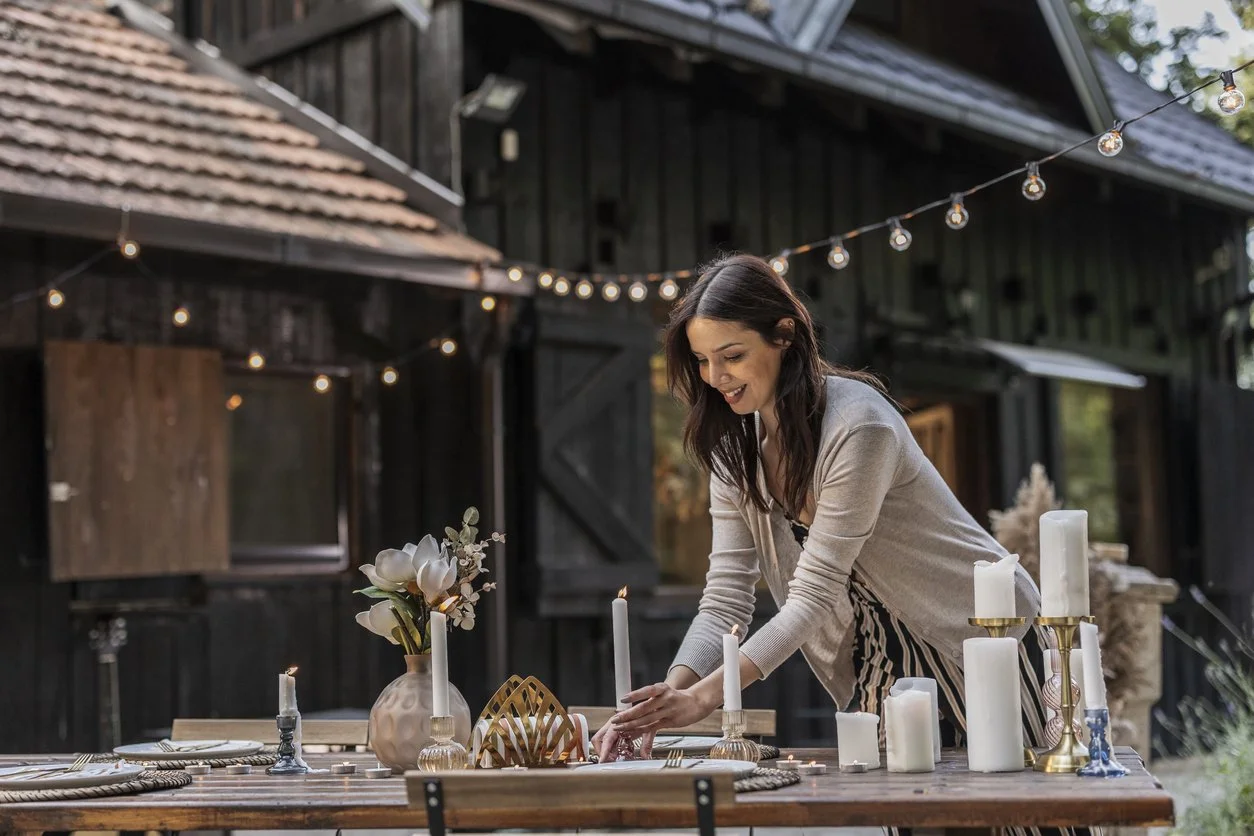 A woman arranging candles and flowers on a rustic outdoor dining table decorated with candles, a vase with white flowers, and gold accents, set in a backyard with string lights and a dark wooden barn in the background.