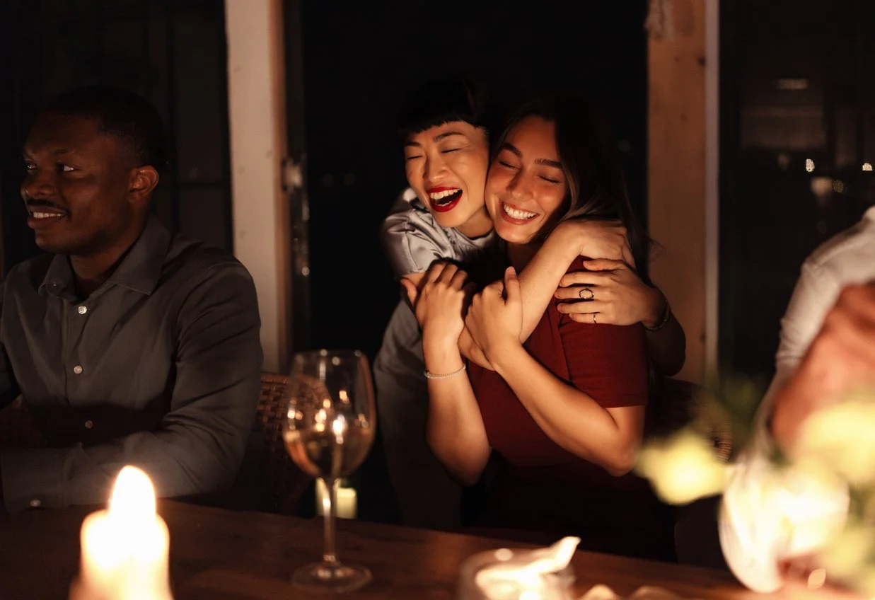 Two women hugging and smiling at a dinner party, candlelight on the table, with a man sitting nearby.