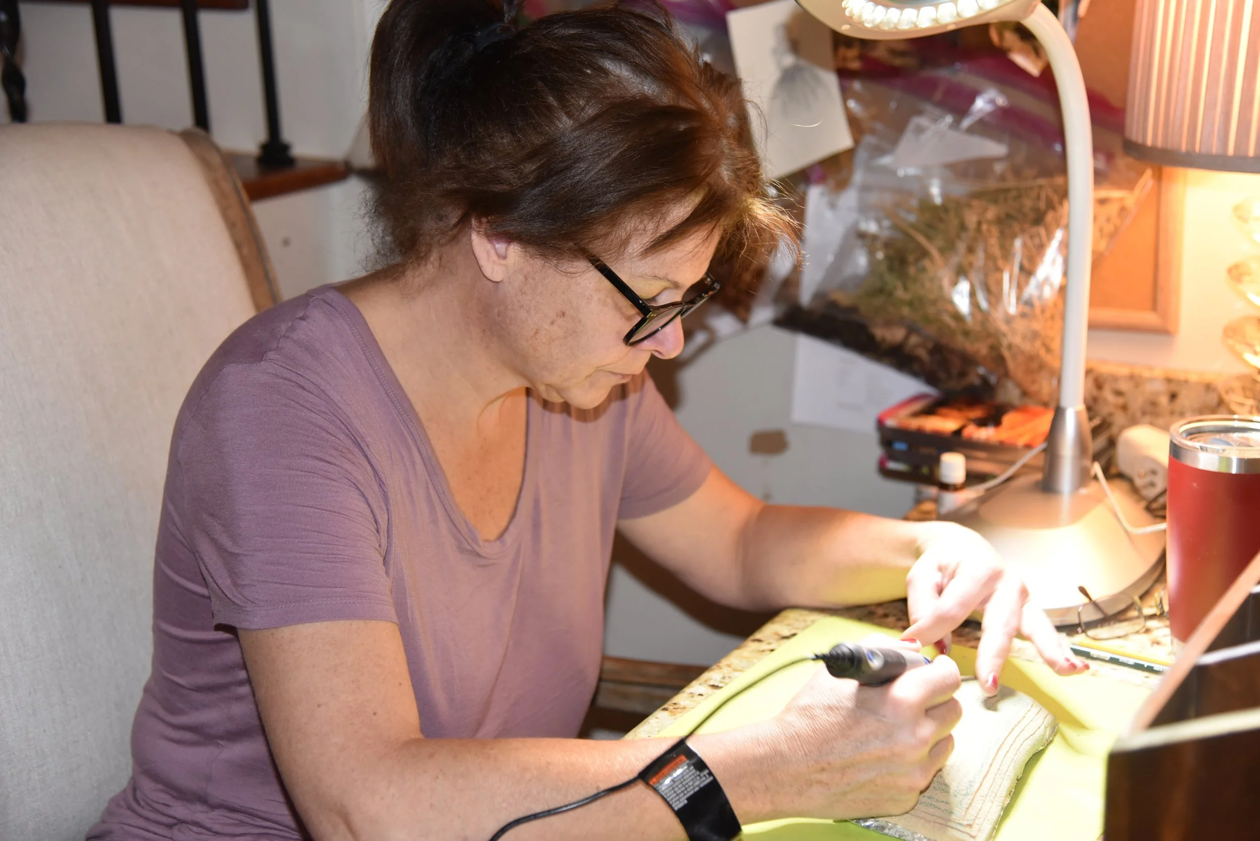 A woman with brown hair and glasses sitting at a desk, focused on a woodworking project, using a rotary tool on a piece of wood, illuminated by a desk lamp.