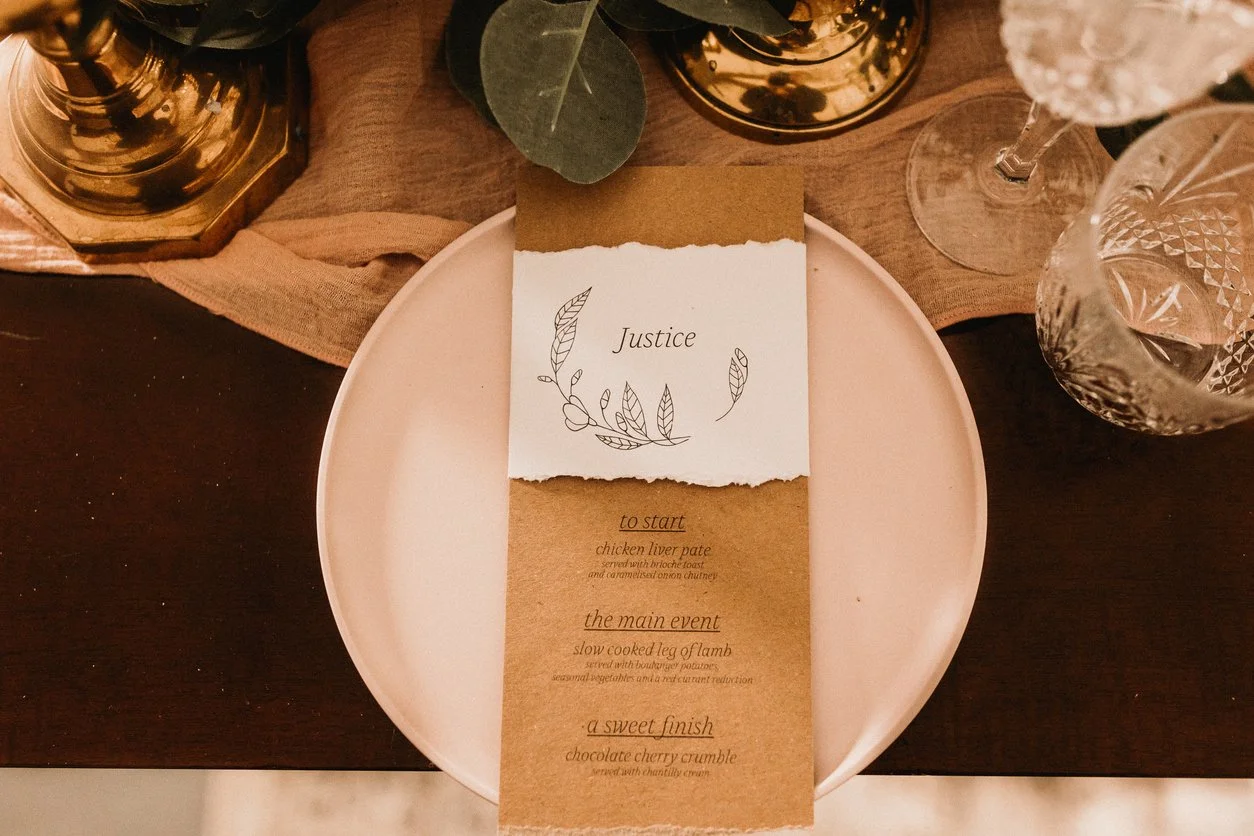 Wedding menu with the title 'Justice', listing three courses: starter chicken liver pate, main slow cooked leg of lamb, and dessert chocolate cherry crumble, placed on a pink plate with glasses and greenery around.