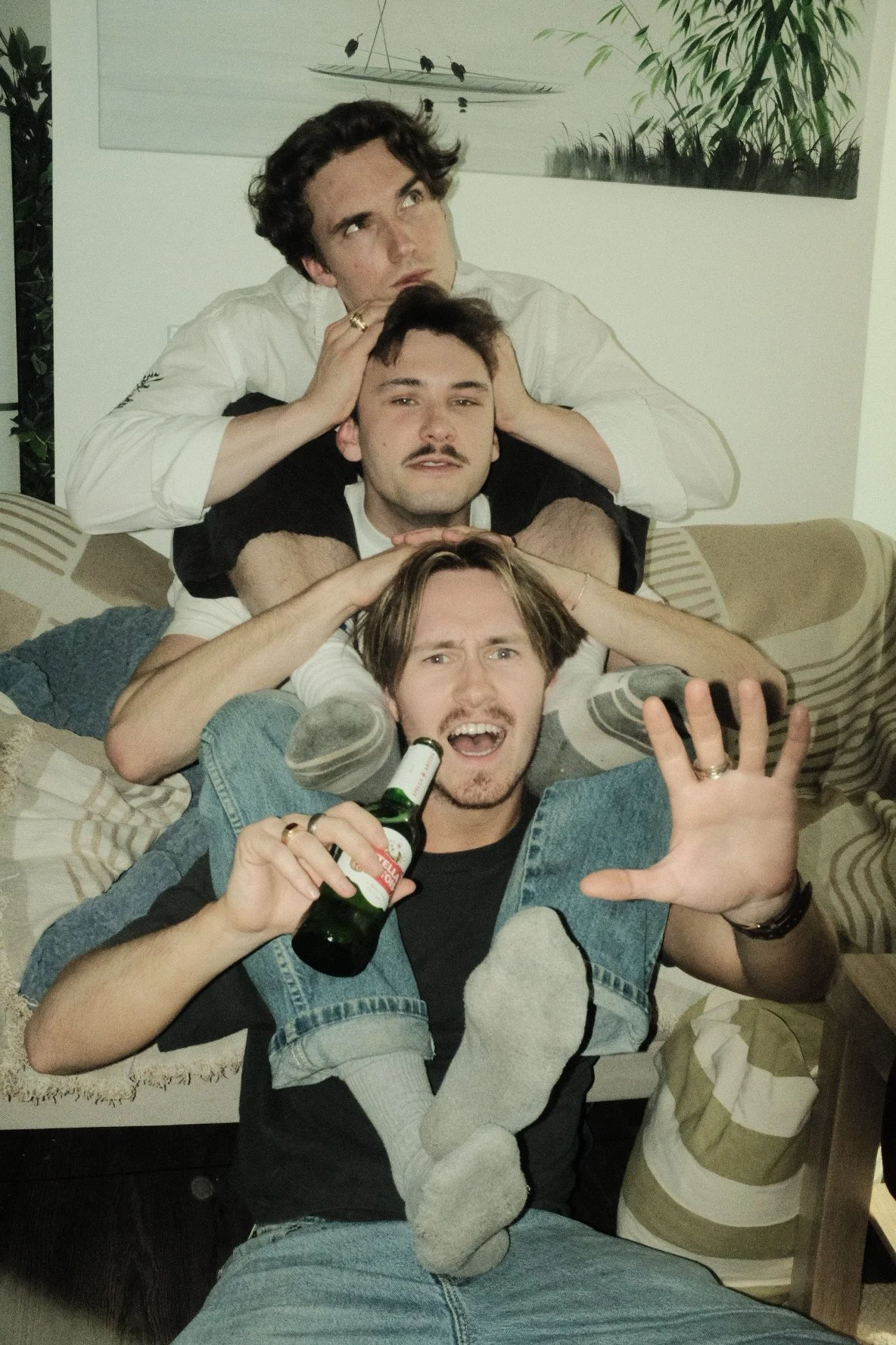 Four young men lying on a sofa in a stack, holding each other's heads and shoulders, with the man at the bottom holding a beer and making a peace sign.