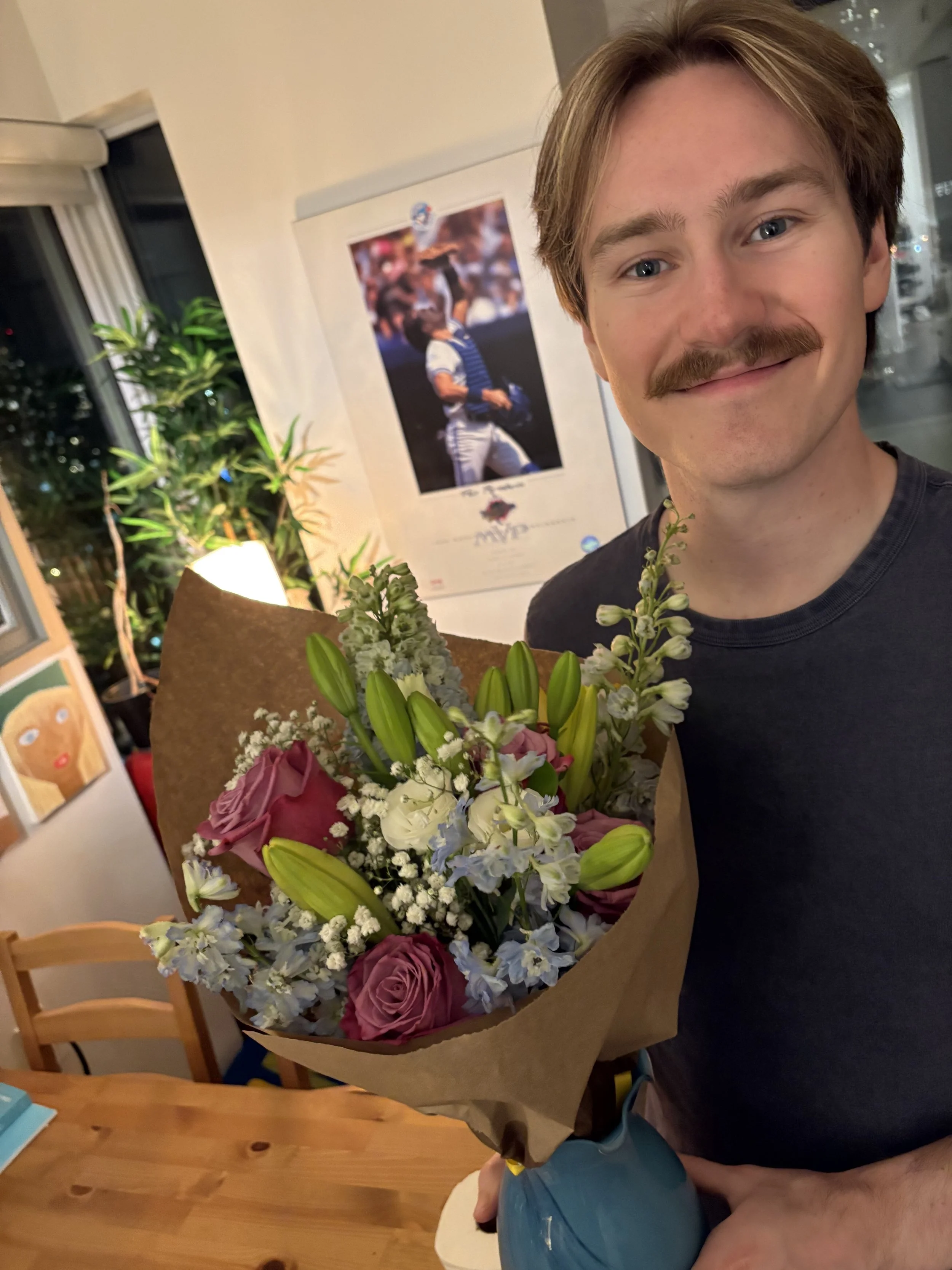 A man with light brown hair, blue eyes, and a mustache is smiling while holding a bouquet of flowers in a room with a wooden table and framed pictures on the wall.