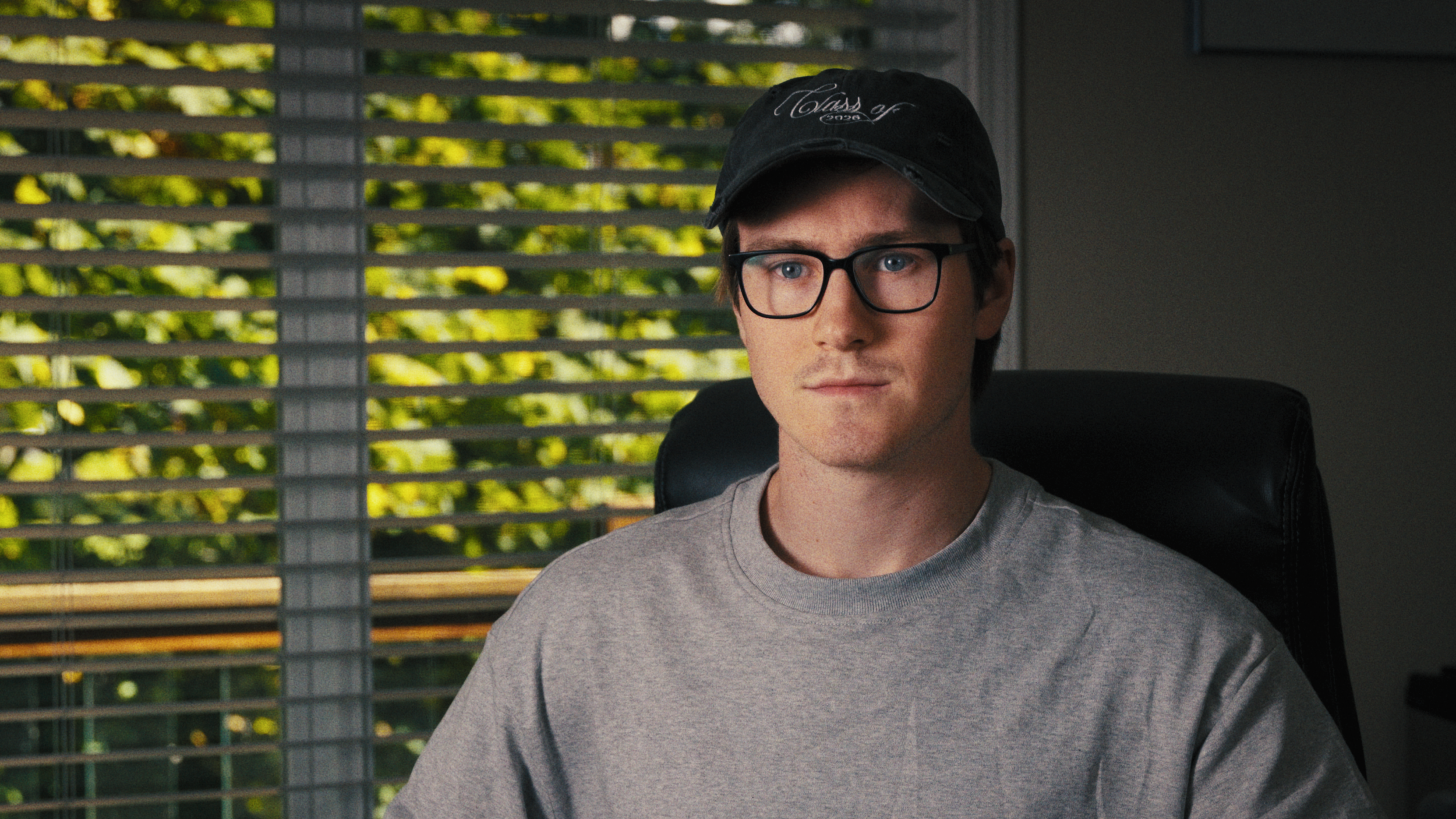 A young man with glasses and wearing a black cap sits in a chair indoors near window blinds, with trees visible outside.