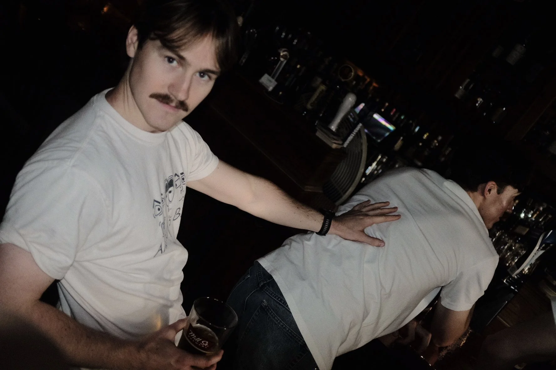 A young man with a mustache, wearing a white t-shirt, holding a dark glass of beer, leaning on the back of a bartender in a dimly lit bar with a shelf of liquor bottles in the background.