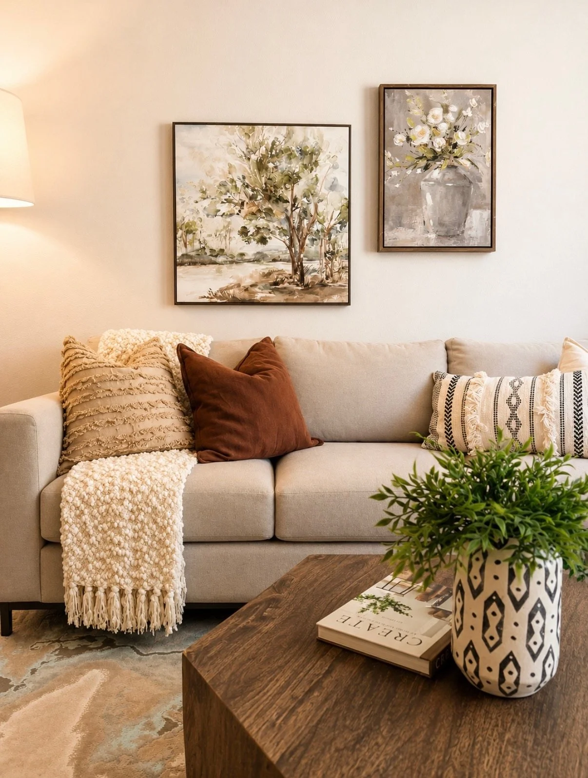 Cozy living room styled with a cream sofa, layered pillows, and throw, wood coffee table, greenery, floral artwork, and a patterned rug.