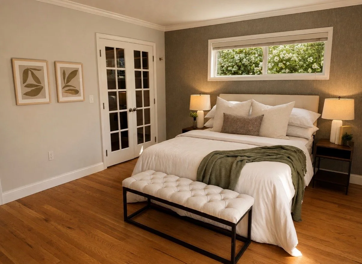 Inviting bedroom staged with layered neutral bedding, a tufted bench, warm wood floors, soft lighting, and a window overlooking greenery.