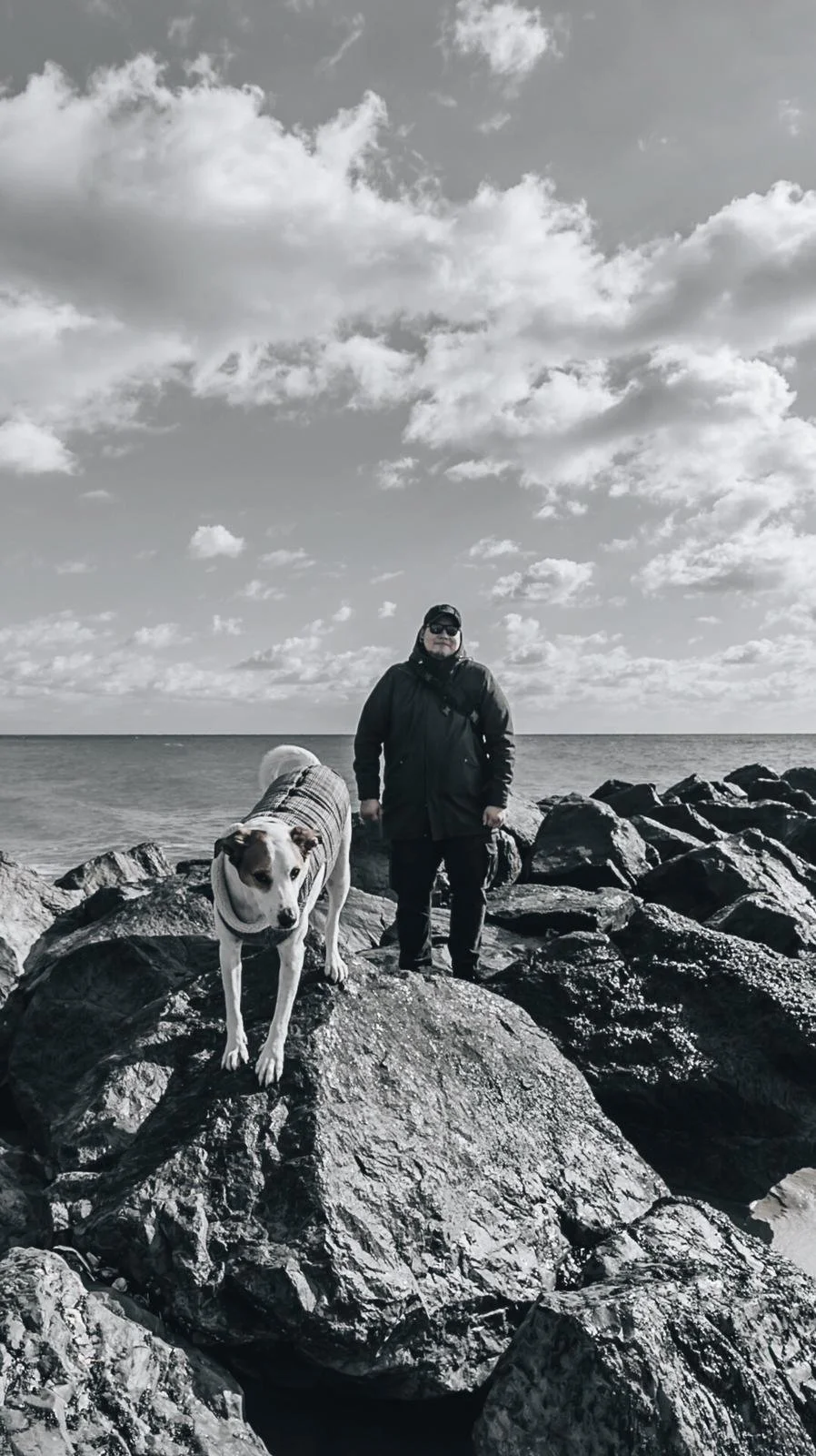 A person wearing dark clothing and sunglasses standing on large rocks at the beach, with a dog draped in a striped sweater nearby, under a cloudy sky.