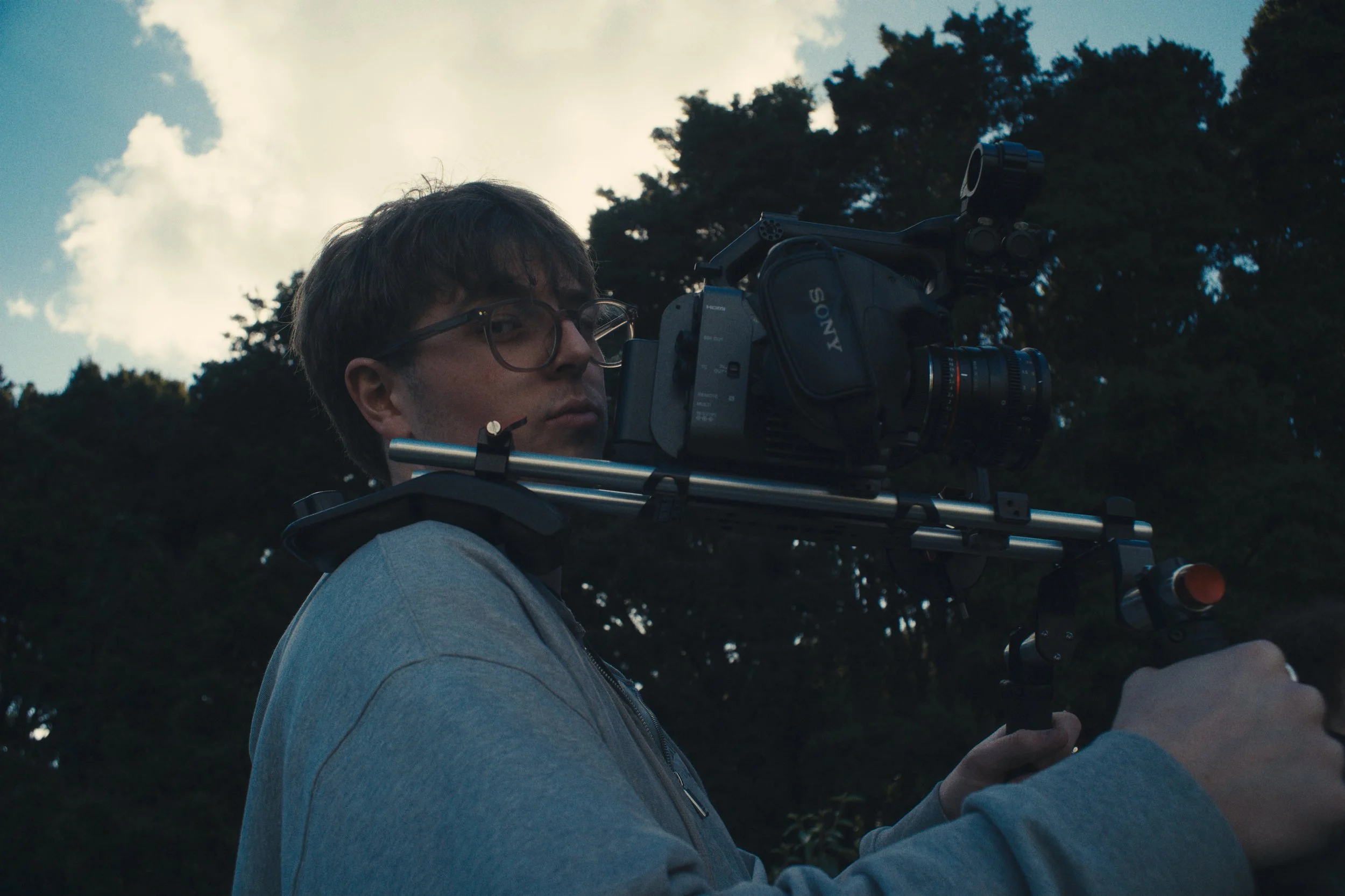 A person with glasses operating a professional video camera outdoors during daytime, with trees and cloudy sky in the background.