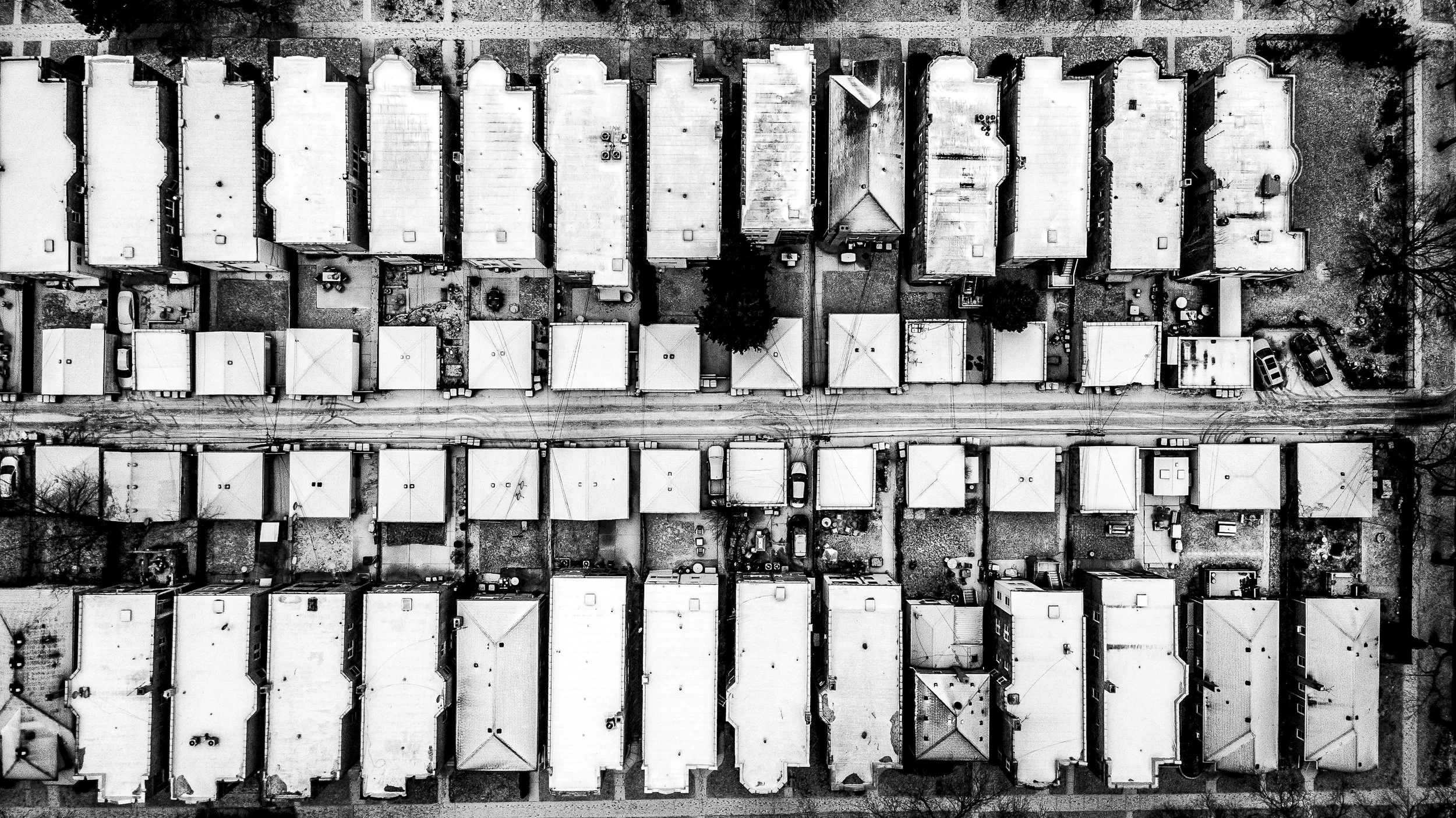 Aerial view of a neighborhood with multiple houses with flat rooftops, cars parked along a street, and trees in between the houses, in black and white.