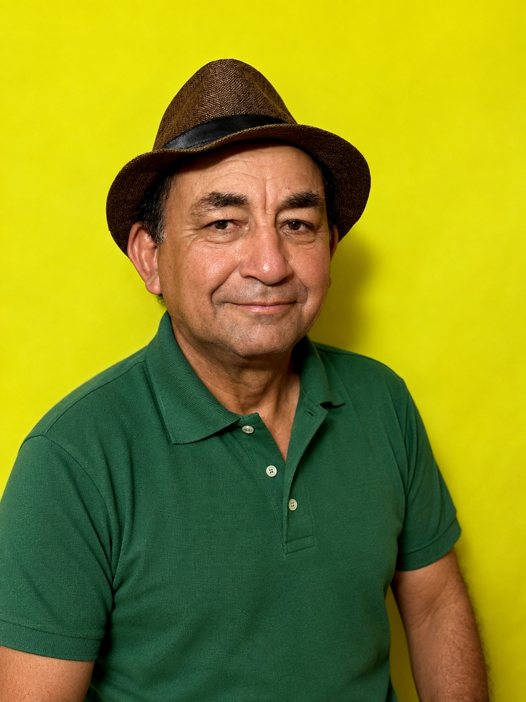 Homem de pele clara, usando camisa polo verde, chapéu marrom com faixa preta, sorrindo, contra fundo amarelo.