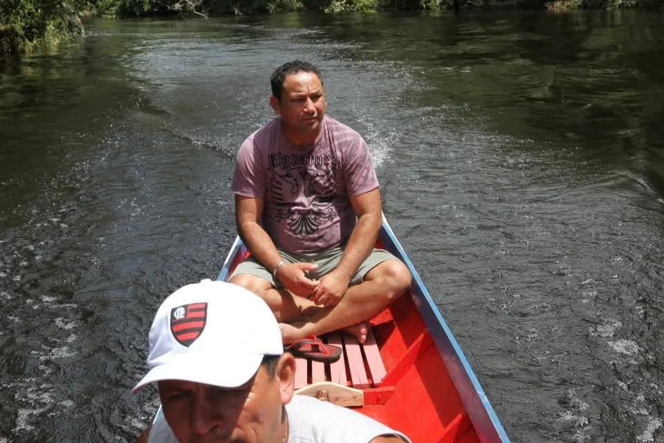 Homem sentado de pernas cruzadas dentro de um barco navegando em um rio, ao fundo há árvores.