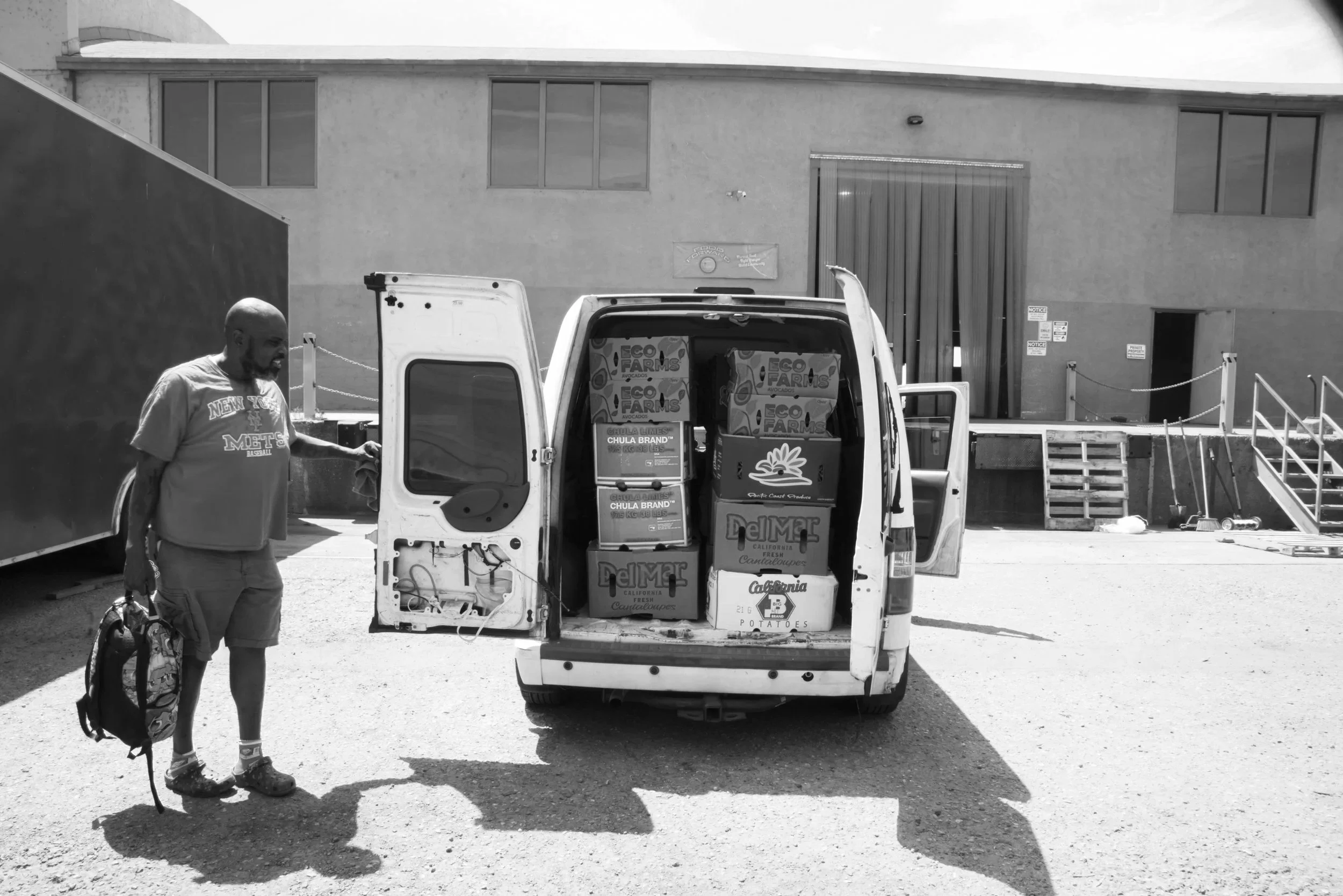 A man holding a bag stands next to a white van with its back doors open, filled with boxes of produce, in an outdoor area with a building in the background.