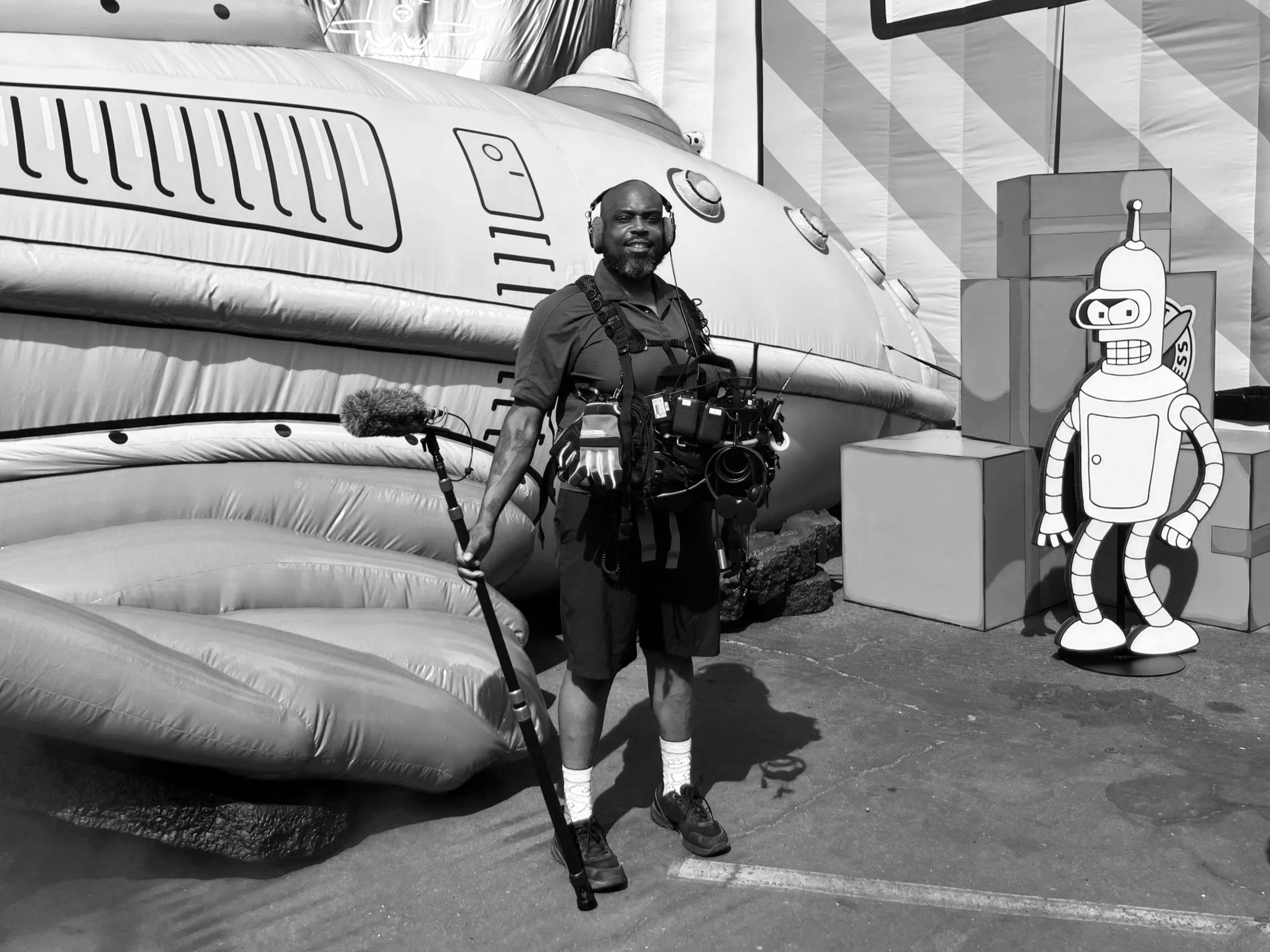 A man with a beard wearing headphones, black shirt, shorts, and sneakers, standing in front of an inflatable airplane prop with cartoon characters on it, at a fair or amusement park.