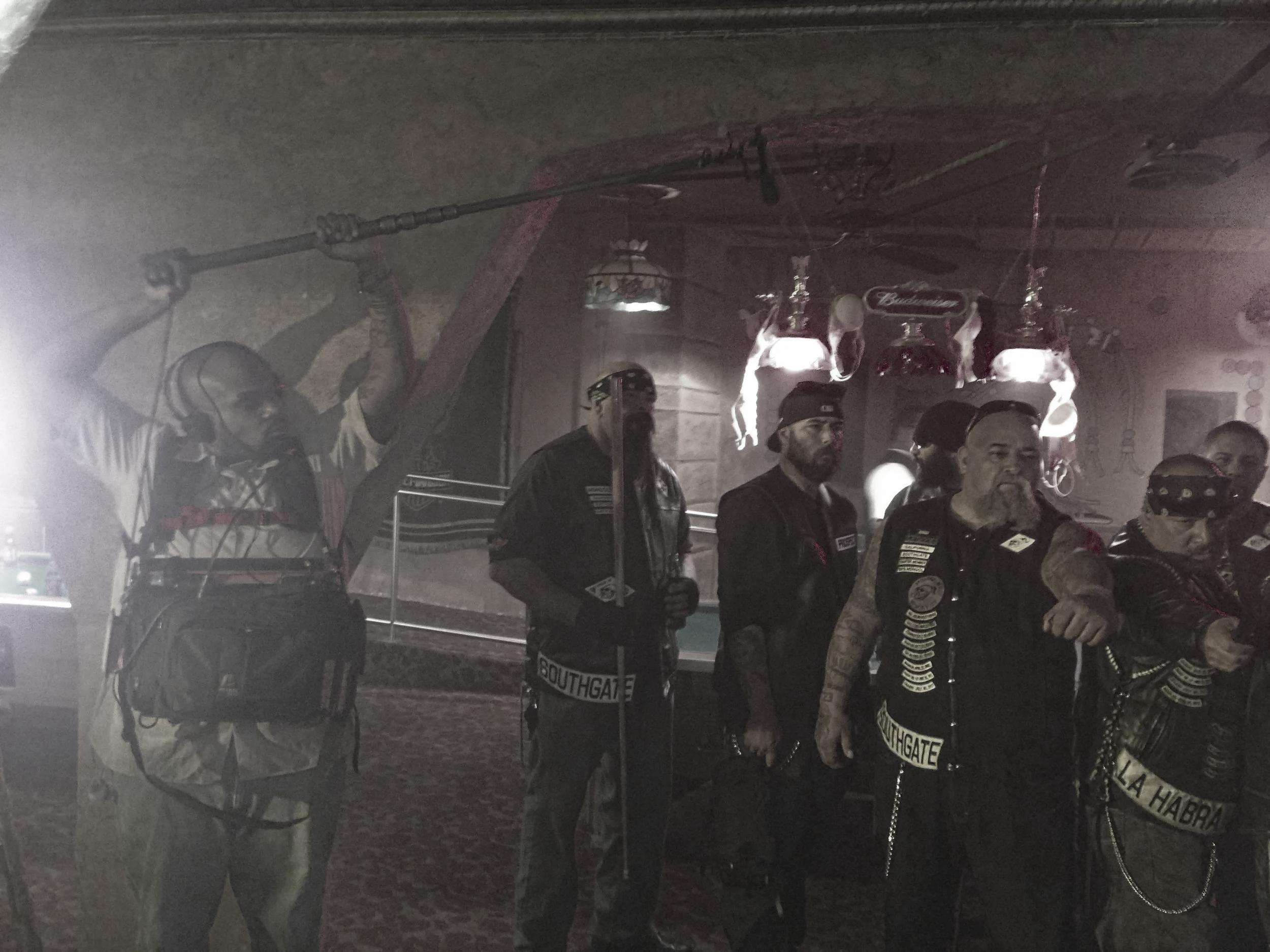 A group of people, some with tattoos, standing indoors at night. One person is holding a drink and wearing a cap. There are hanging lights and some decorations in the background.