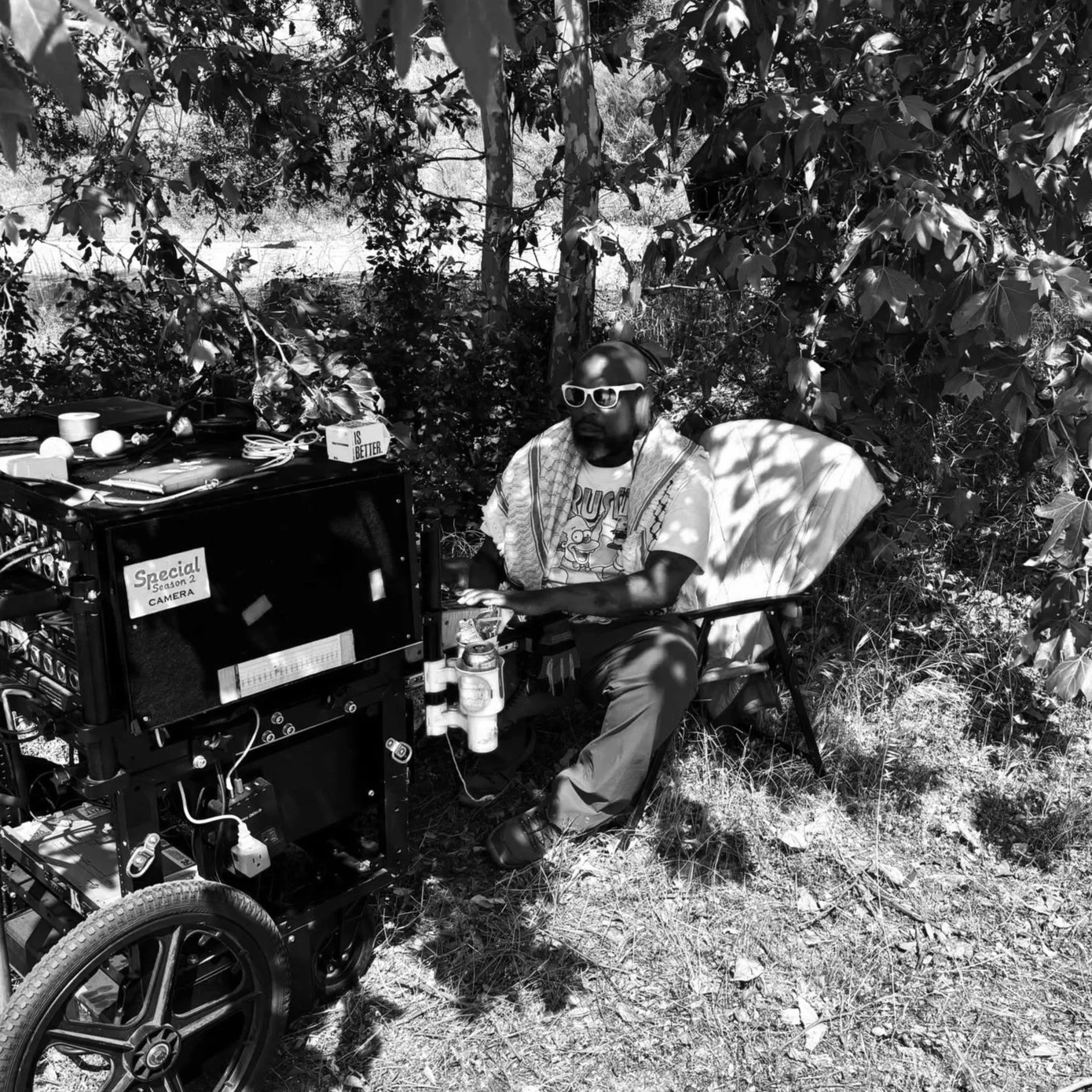 A man sitting outdoors surrounded by trees, wearing sunglasses and headphones, with a towel around his neck, next to a setup of electronic equipment connected to a wheelchair.