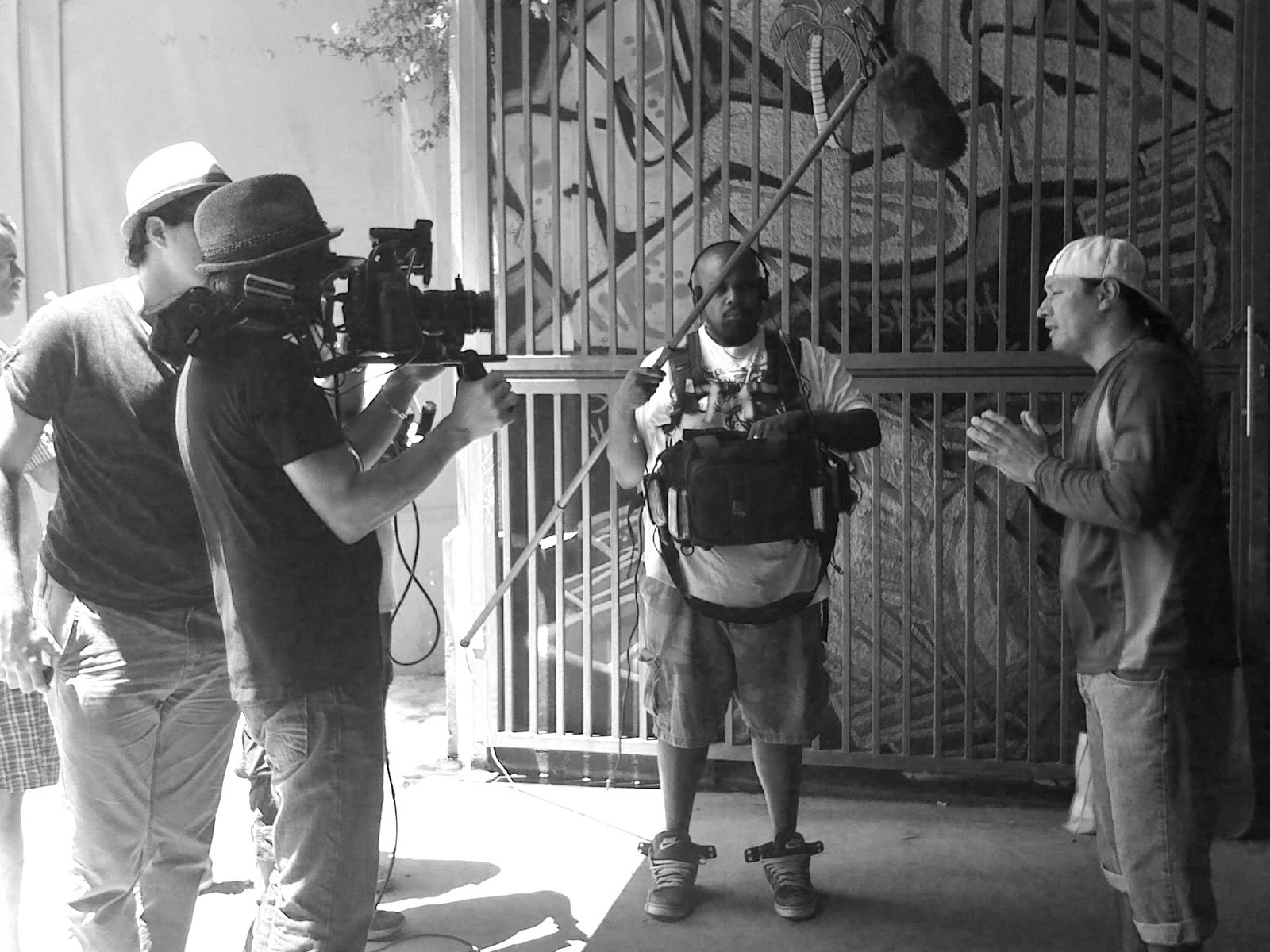A black and white photo of a video shoot or interview happening outdoors. Four men are the focus; one man is speaking to another who is listening, with the third man operating a camera and the fourth standing nearby. The man in the center has a backpack and a pole, possibly microphone or lighting equipment.