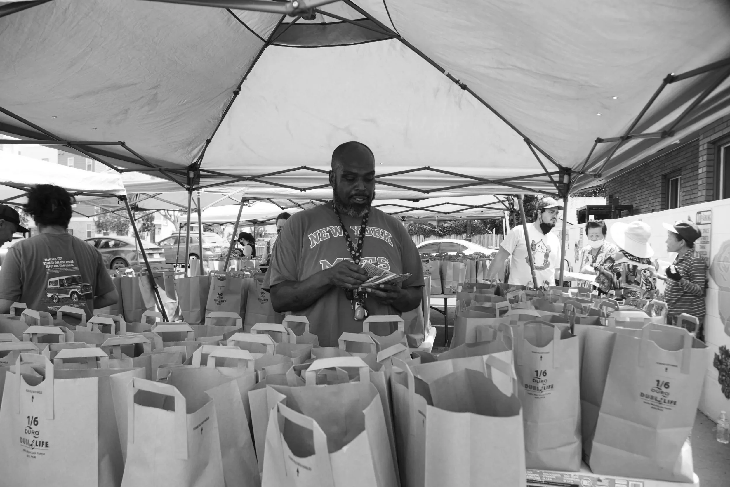 A man in a hoodie reading a booklet at an outdoor market stall with paper bags on display, under tents, with other people browsing and buildings in the background.