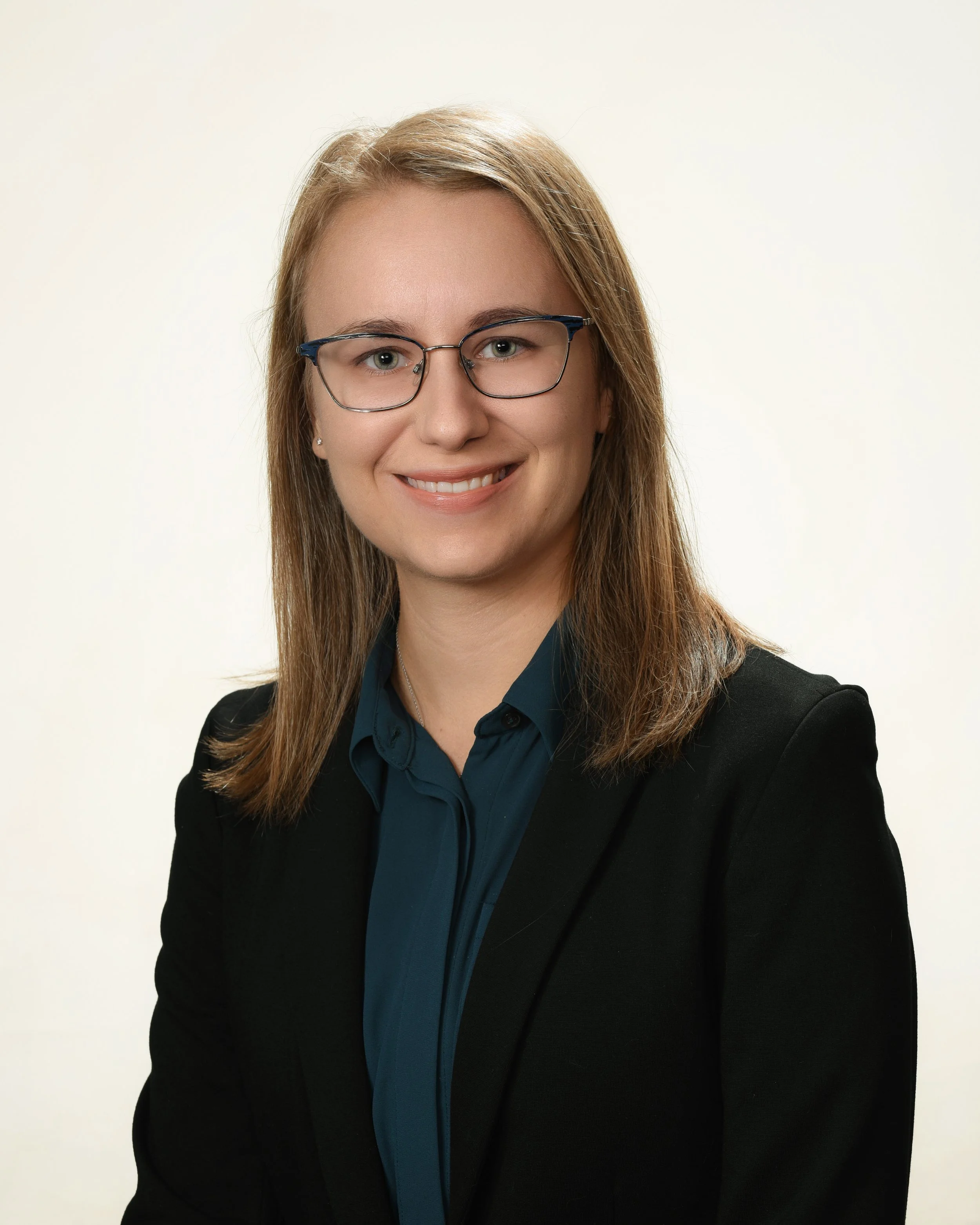 A woman with long brown hair, glasses, and a bright smile, wearing a dark blazer and a blue shirt, posed against a plain white background.