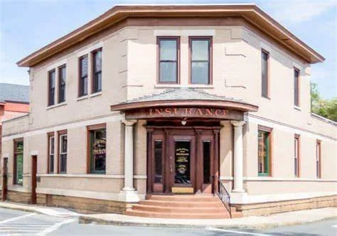Two-story beige building with an insurance sign above the entrance, featuring wooden doors, columns, and multiple windows.
