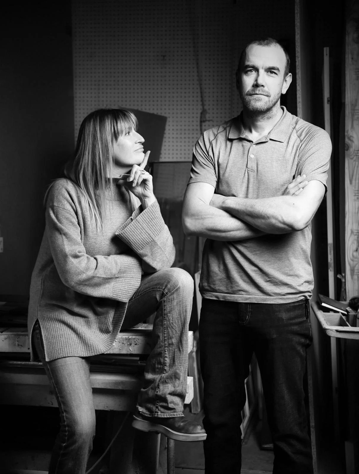 A black and white photo of a man and woman in a workshop or studio. The woman is sitting on a workbench, looking thoughtfully at the man. The man is standing with arms crossed, looking directly at the camera. The background includes pegboard walls and construction materials.