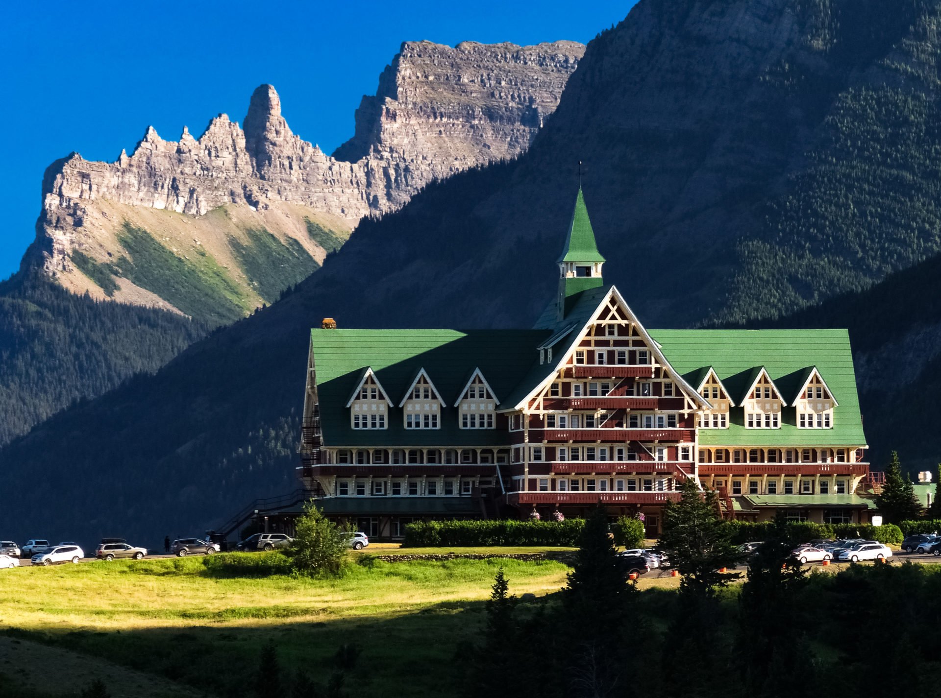 A large hotel with a green roof and multiple balconies, set against a backdrop of tall mountains with rocky formations and lush green forests. There are parked cars and a grassy area in the foreground.