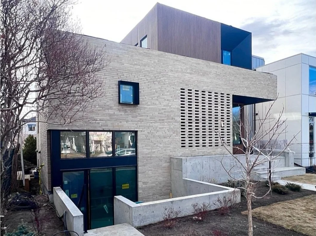 Modern multi-story residential building with a beige brick facade, large windows, and a concrete entrance staircase, set in a landscaped yard with leafless trees.