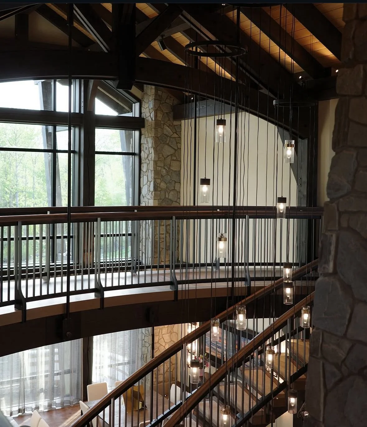Interior of a multi-story building featuring large windows, a curved wooden staircase with black metal railings, and hanging pendant lights with exposed bulbs.